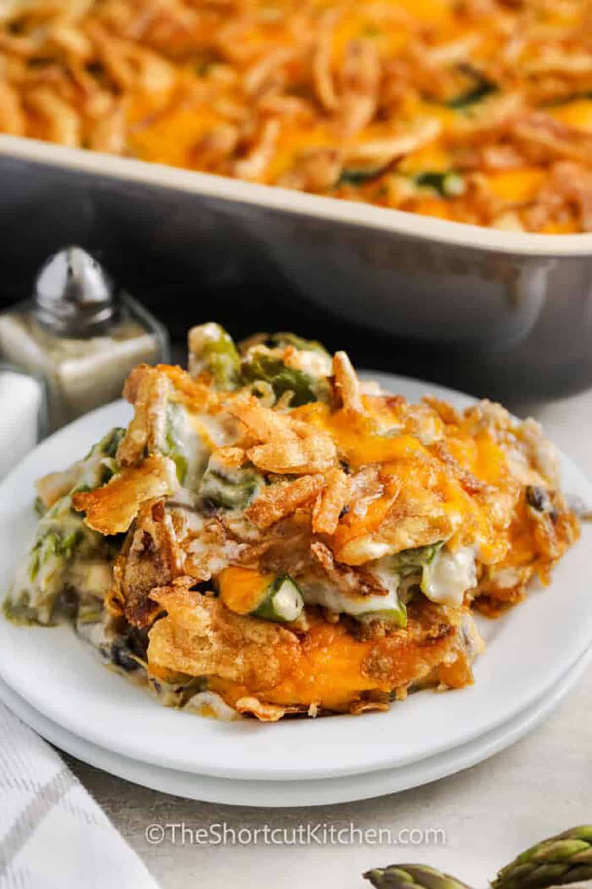 Asparagus casserole recipe is a creamy, cheesy side dish that’s packed with flavor and comfort. Tender asparagus is mixed with a rich mushroom sauce, gooey cheddar cheese, and crispy fried onions for the perfect balance of textures. It’s an easy way to turn a simple vegetable into a dish everyone will love. Whether for a holiday meal or a weeknight dinner, this cheesy bake is always a hit! #theshortcutkitchen plated cheesy Asparagus Casserole Recipe