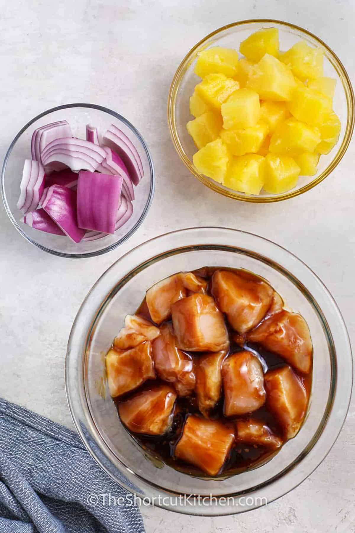 Chicken pineapple kabobs are a sweet and savory grilled dinner that keeps things easy with bottled teriyaki sauce and canned pineapple chunks. Thread chicken, pineapple, and red onion onto skewers, grill until the chicken is juicy, and brush with extra sauce at the end. This shortcut chicken skewer recipe is perfect for warm-weather meals and cookouts. Save this recipe when you need an easy chicken dinner. #theshortcutkitchen marinated chicken and a bowl of onions and pineapple to make Chicken Pineapple Kabobs