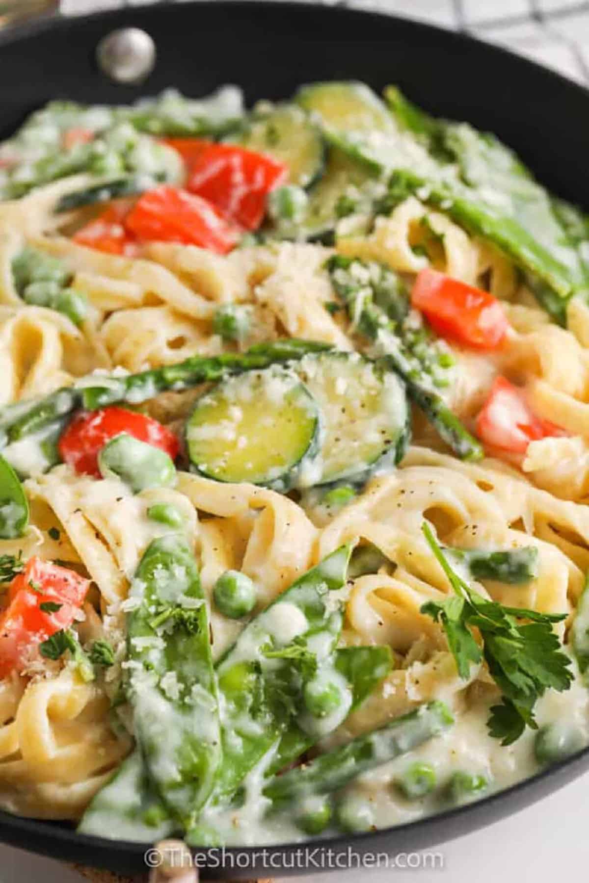 Looking for a colorful, light, and creamy dinner? This recipe for pasta primavera is the answer! With fresh spring veggies like peas and zucchini, this easy one-pot meal is full of flavor. The creamy Alfredo sauce ties everything together, making it a perfect meatless weeknight dinner. The vibrant vegetables are sautéed in butter and garlic, then simmered in the sauce before being tossed with pasta. You’ll love how simple and satisfying this dish is, ideal for a busy evening. #theshortcutkitchen close up of Pasta Primavera with parmesan in a pan