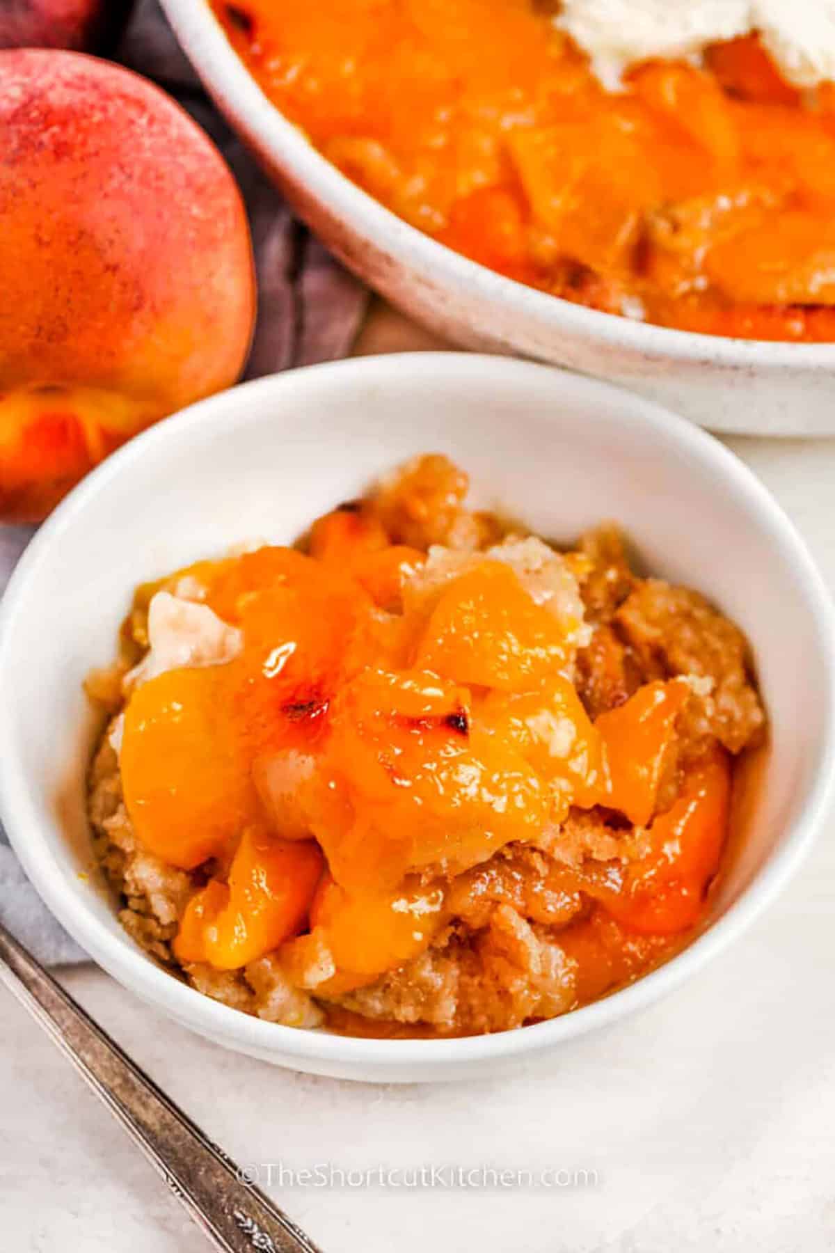 This Bisquick peach cobbler recipe has rich, homemade flavor in a quick and easy dessert. Using shortcut ingredients like biscuit mix and canned peaches, this recipe comes together fast and bakes up golden, fluffy, and sweet. Peach cobbler made with Bisquick is a Southern classic and is always welcome at summer picnics and barbecues. Serve it warm with a scoop of vanilla ice cream for a mouthwatering treat that no one can resist. #theshortcutkitchen plated Bisquick Peach Cobbler Recipe