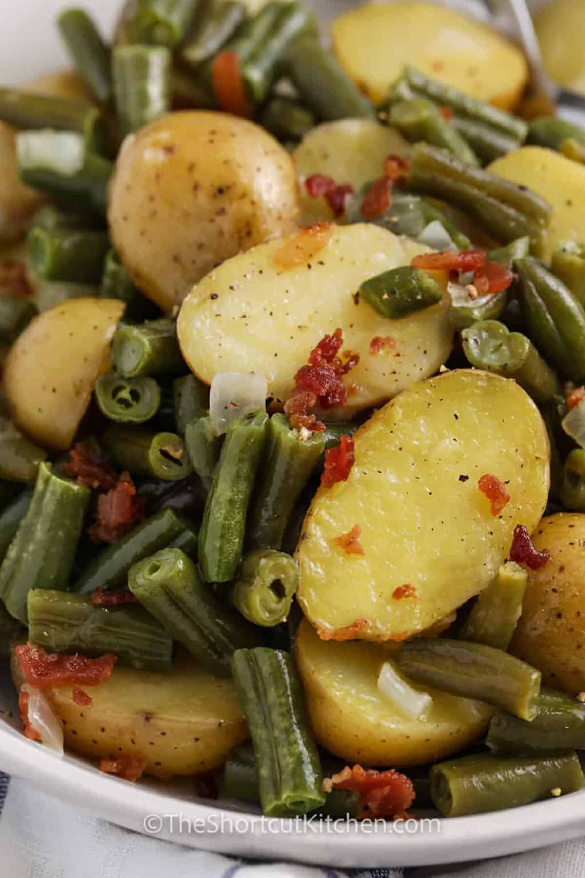 Southern green beans with potatoes is an old-fashioned, country style favorite made easy in one pot. Baby potatoes and crisp green beans are simmered in savory chicken broth, sautéed with onion and garlic for depth, and seasoned perfectly. Smoky bacon adds richness to every bite. This hearty side is pure comfort food, perfect for weeknights or a Sunday supper spread. #theshortcutkitchen bowl of Green Beans With Potatoes