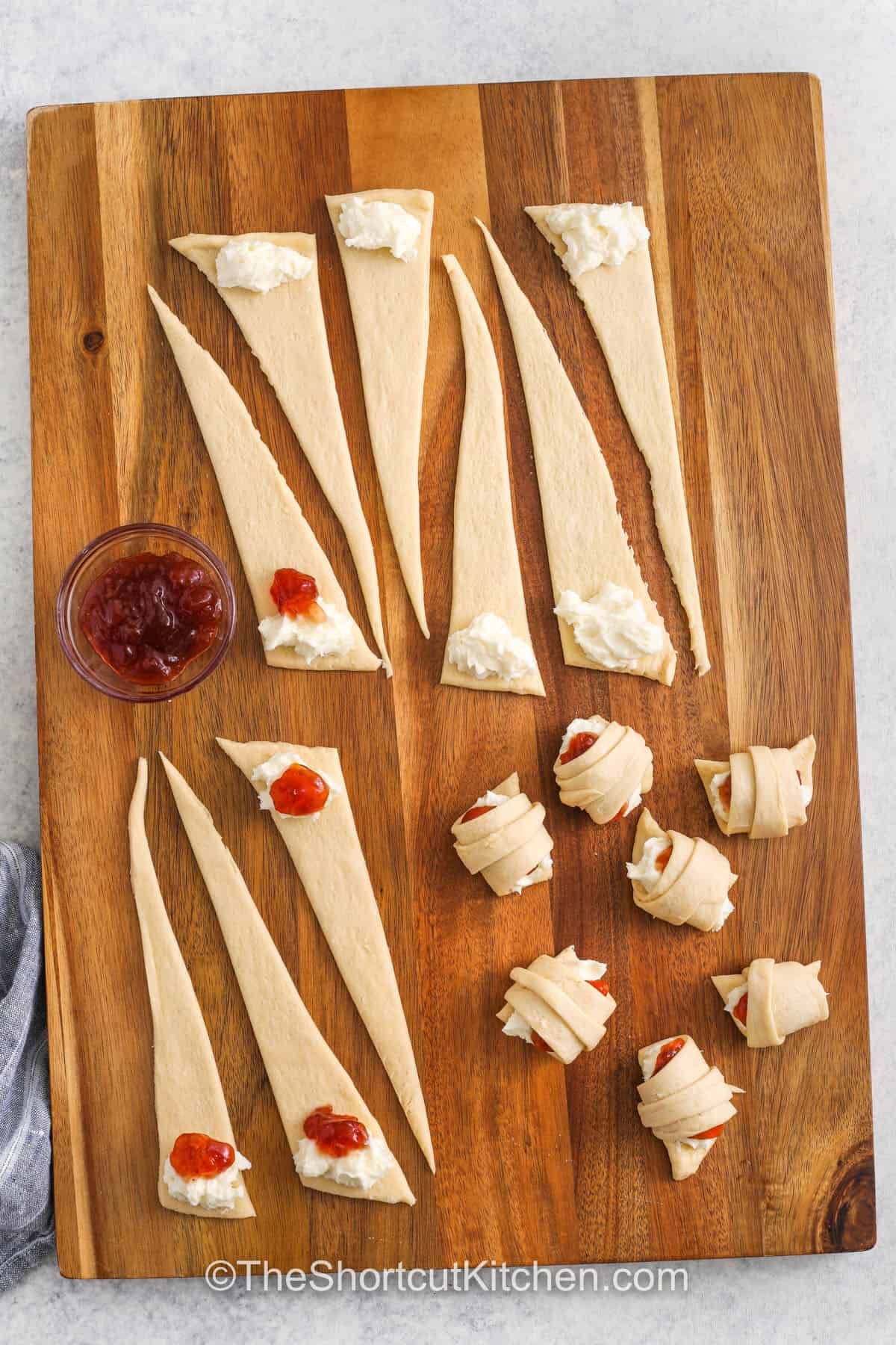 Cream cheese crescent rolls are the perfect combination of sweet, fruity, and creamy, all in one easy breakfast pastry. This recipe starts with crescent dough filled with a smooth cream cheese mixture and fruit jam, either cherry, apple, blueberry, or strawberry. Brush with butter and bake until golden, then sprinkle with cinnamon or finish with chocolate for a bakery-style dessert. These homemade danishes bake up buttery and fluffy and taste amazing! #theshortcutkitchen adding cream cheese and jam to crescent roll dough and rolling up to make Cream Cheese Crescent Rolls