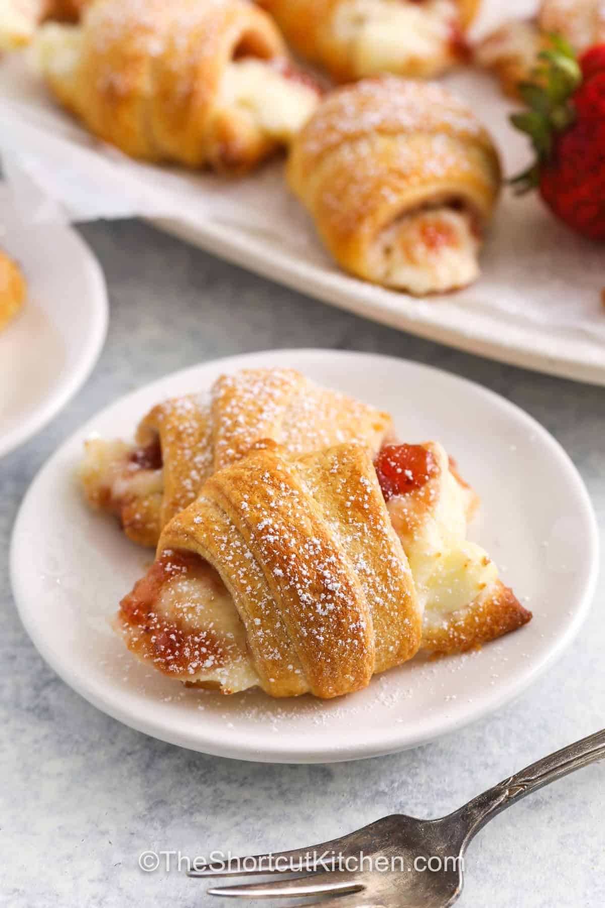 This cream cheese crescent rolls recipe makes an easy and delicious pastry that’s perfect for dessert or a sweet snack. Flaky crescent roll dough is filled with sweetened cream cheese and your favorite fruit jam or pie filling. Use cherry, blueberry, apple, or strawberry. Sprinkle with cinnamon or drizzle with chocolate for a bakery-style danish. It’s a fun recipe to make and even better to eat! #theshortcutkitchen Cream Cheese Crescent Rolls on a plate