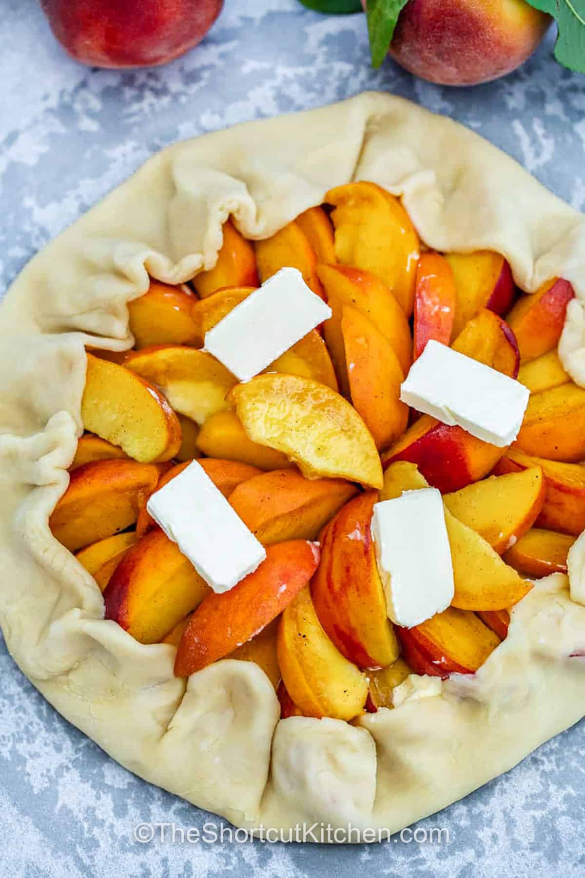 A good peach galette recipe should be simple and full of fresh flavor, and this one nails it. With a homemade feel and rustic look, it layers tart-sweet peaches inside a buttery crust that turns golden in the oven. The open-face style is so easy to shape and perfect for showing off those juicy summer peaches. Sprinkle the edges with cinnamon sugar and serve with ice cream for a treat everyone will remember. #theshortcutkitchen folding pastry dough over peach mixture to make Peach Galette Recipe