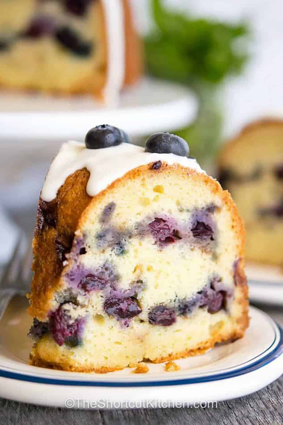 Blueberry sour cream coffee cake is an easy recipe that tastes just like something from a bakery. It’s got a sweet cinnamon swirl, a soft, moist crumb, and juicy blueberries in every bite. This old fashioned favorite has a no fuss method and a simple glaze topping that makes it look as good as it tastes. Perfect as a make ahead treat for brunches or coffee breaks. Serve it with coffee or pack it for a sweet mid-day snack. It’s just right for sharing. #theshortcutkitchen close up of a piece of Blueberry Coffee Cake Recipe