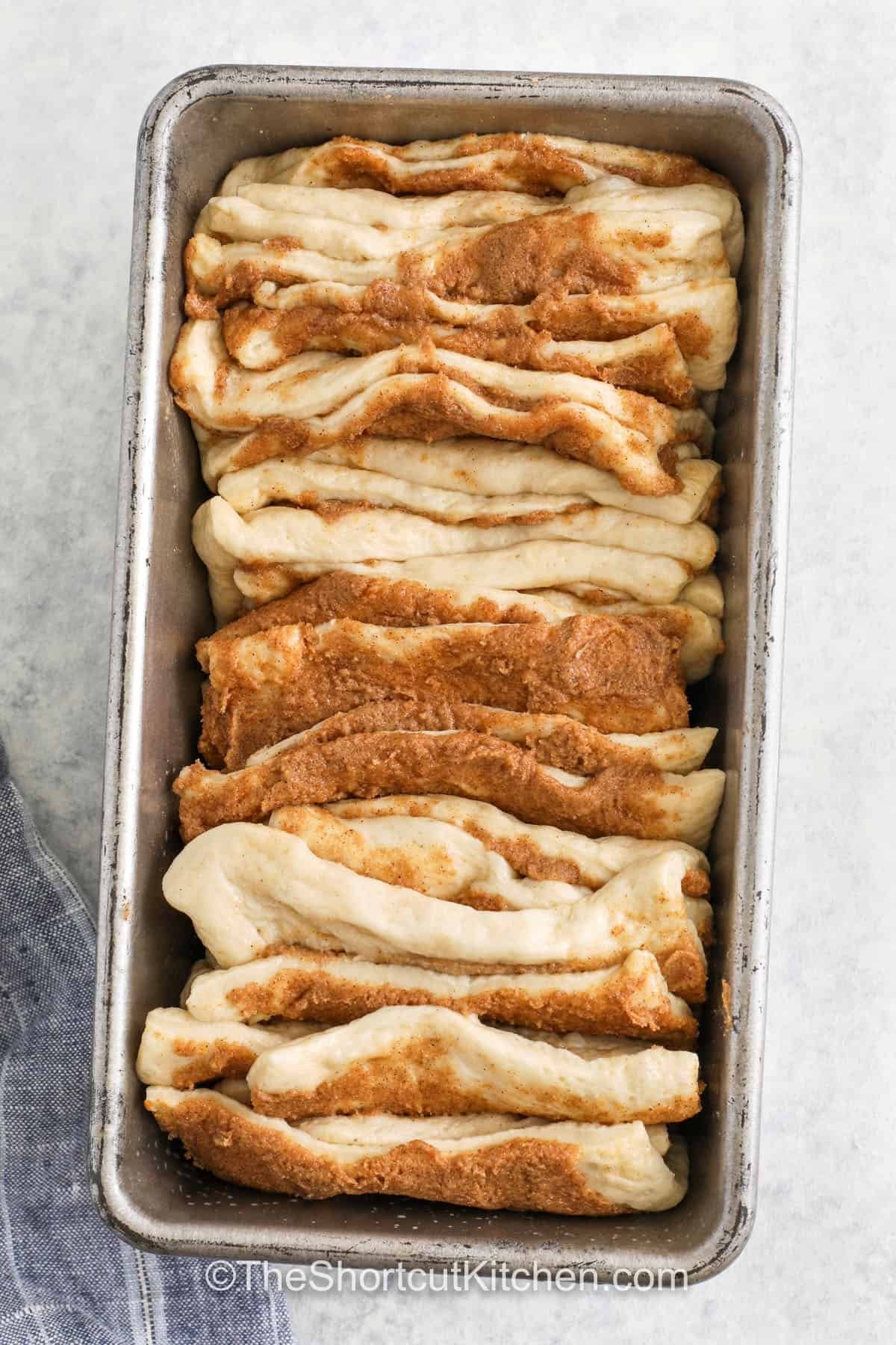 Enjoy a homemade treat the easy way with this simple cinnamon pull apart bread! Refrigerated pizza dough is sliced and stacked into easy pull-apart layers with a buttery cinnamon-sugar filling in between. After baking to a toasty golden brown, give it a drizzle of sweet vanilla glaze to produce a fun version of cinnamon monkey bread. All you have to do next is gather the family around and watch it disappear! #theshortcutkitchen Cinnamon Pull Apart Bread in the dish before baking
