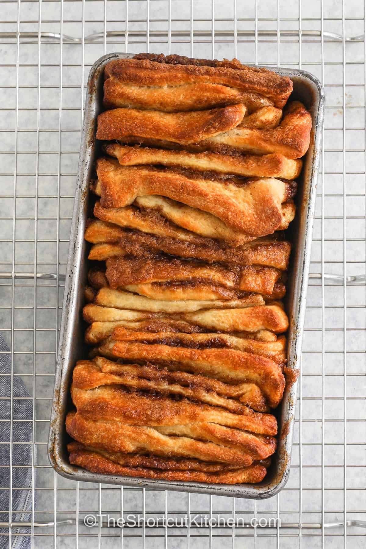If you enjoy monkey bread, you’ll swoon for this easy cinnamon pull apart bread! Start by rolling out refrigerated pizza dough and spread with a mixture of butter, brown sugar, and cinnamon. Then slice, stack into layers, and bake it until it fluffy, and golden. Each sticky layer is delicious unfrosted, but you can drizzle it with this sweet vanilla glaze for extra dose of yum. Customize and vary the flavors however you want. Try it with apples, chocolate, or pumpkin spice. #theshortcutkitchen Cinnamon Pull Apart Bread in the dish on a cooling rack
