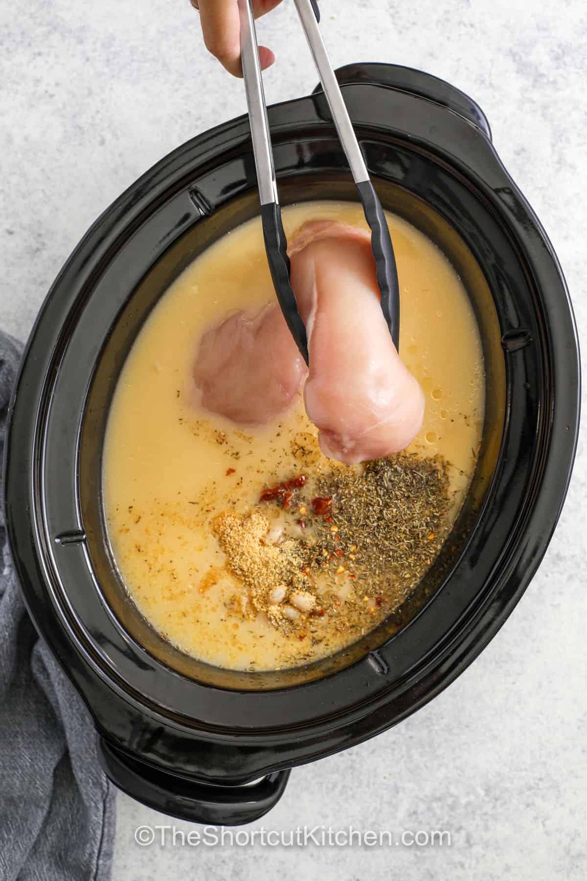 Tuscan chicken soup is creamy, hearty, and full of comforting flavor. The slow cooker is perfect for cooking, blending the flavors and textures of shredded chicken, white beans, spinach, and sun-dried tomatoes into a rich broth with garlic and Parmesan. This easy recipe is perfect for a cozy dinner, and pairs beautifully with a slice of crusty bread for dipping. #theshortcutkitchen adding chicken to crockpot to make Crockpot Tuscan Chicken Soup