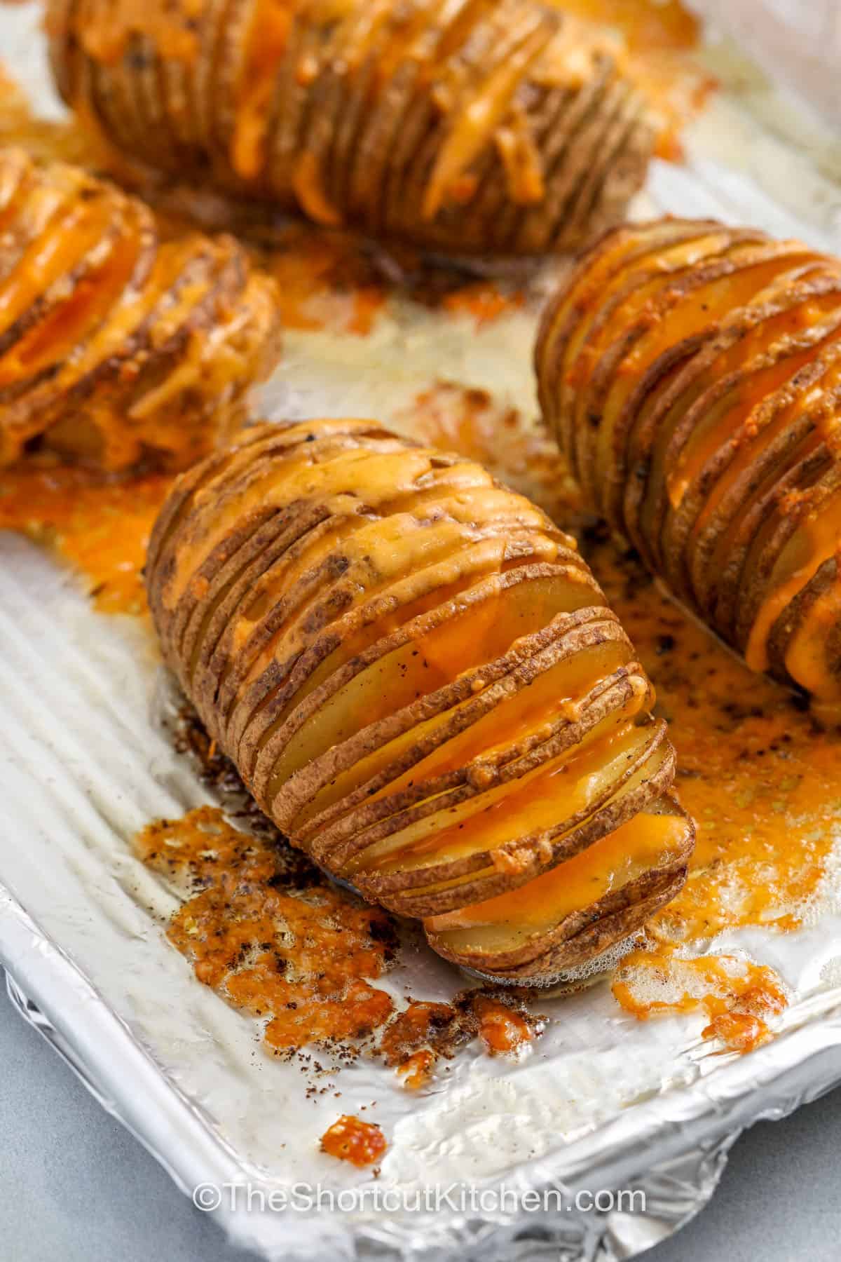 Make your dinner shine with this easy Hasselback potatoes recipe, full of crispy edges and cheesy flavor. Russet potatoes are sliced thin, layered with garlic butter, and roasted in the oven until tender inside and crunchy outside. Sprinkle bacon and cheese in between the slices for a holiday-worthy side dish. It’s the best way to turn a simple potato into something special. #theshortcutkitchen. close up of Hasselback Potatoes Recipe made with cheese on a sheet pan
