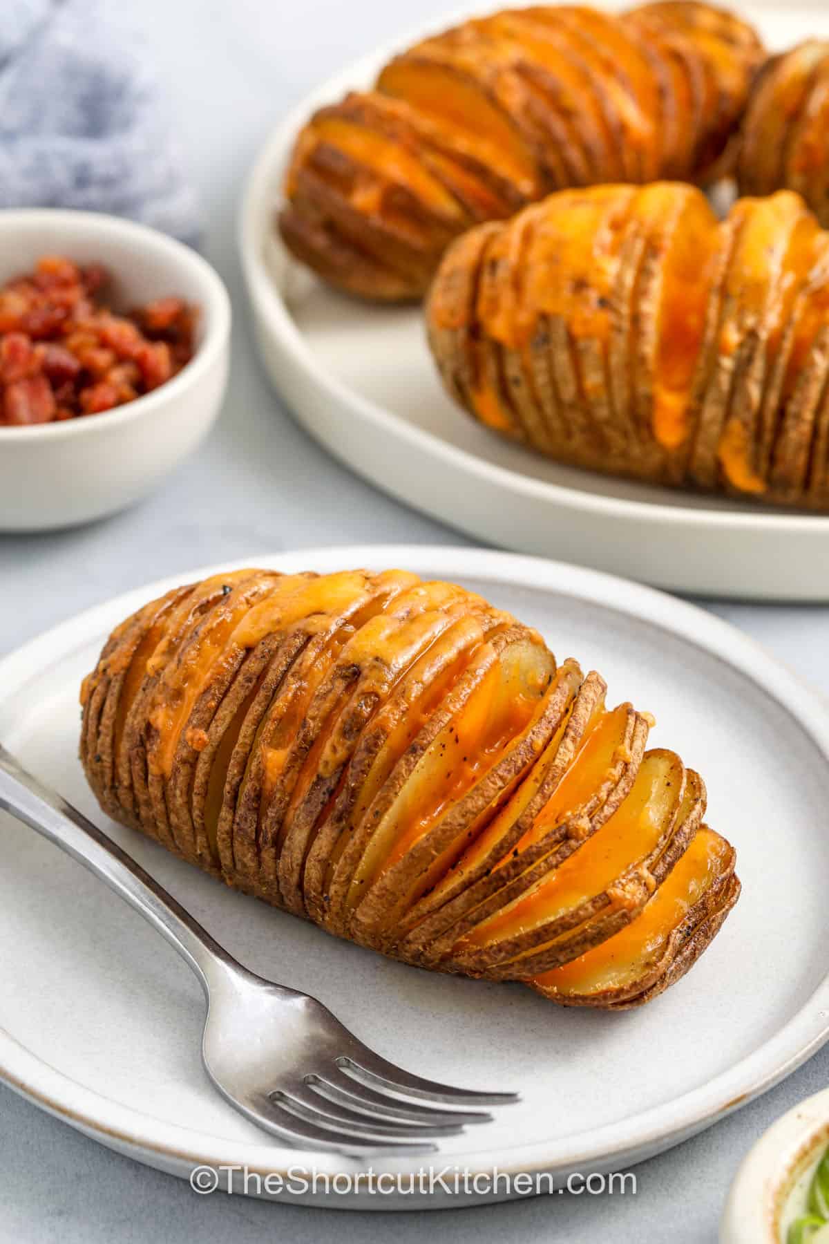This easy Hasselback potatoes recipe makes the best cheesy and crispy side for any dinner. Each potato is thinly sliced, brushed with garlic butter, and baked in the oven until golden. Add bacon and cheese between the slices for a melty surprise that makes this dish perfect for a holiday table. These potatoes look fancy but are simple enough to whip up any night of the week. #theshortcutkitchen. plated Hasselback Potatoes Recipe with a fork