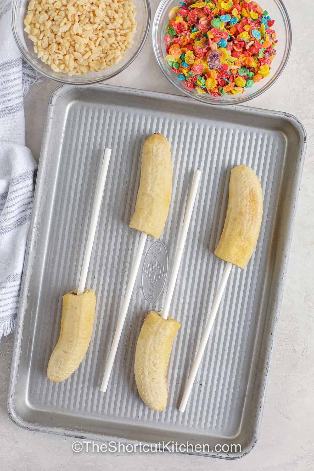 Banana pops are a fun and tasty way to enjoy fruit. These frozen treats are dipped in creamy yogurt and covered in your favorite toppings like cereal, nuts, or coconut. They make an easy recipe for a quick summer snack that is both kid-friendly and delicious. Keep a batch ready in the freezer for a cool and simple treat everyone can grab and enjoy. Perfect for sunny afternoons or anytime you want something light. #theshortcutkitchen frozen bananas on a stick on a baking sheet
