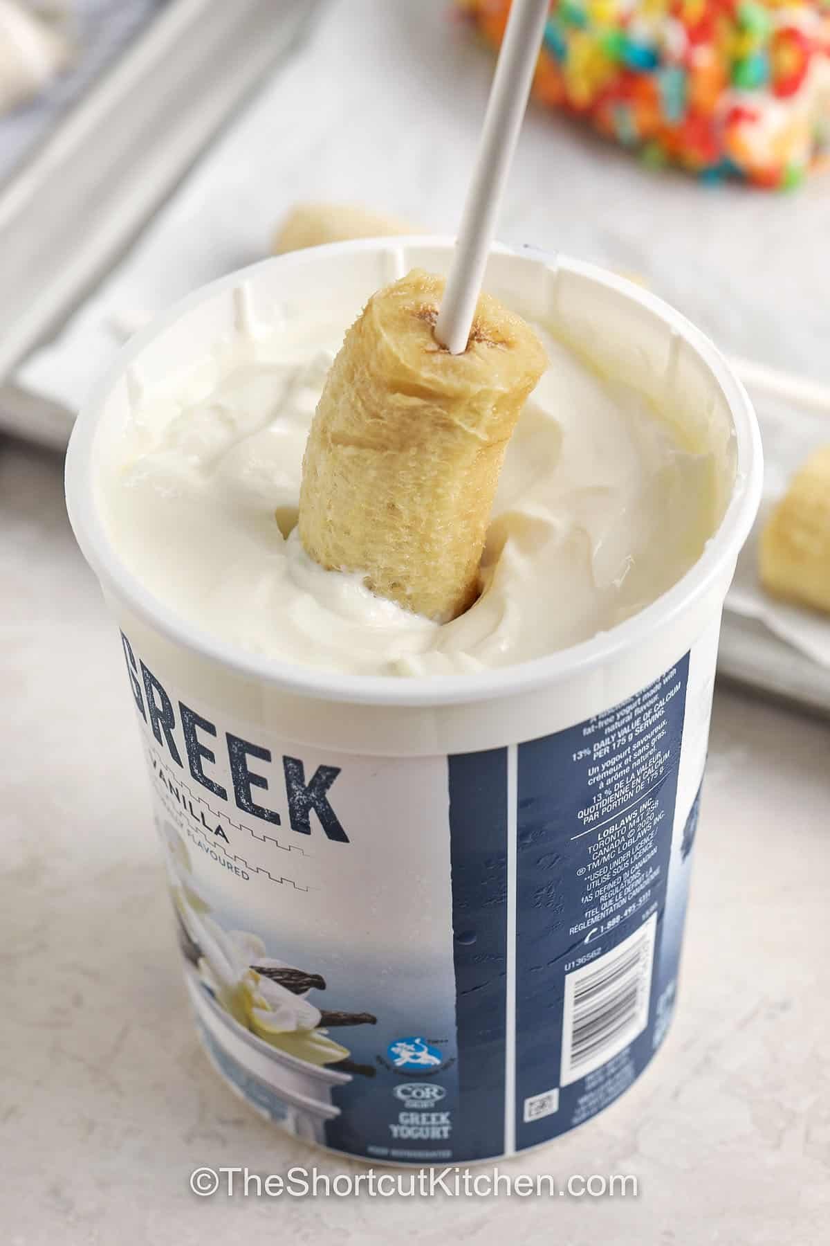 Banana pops are dipped, covered, and frozen for the ultimate snack. This easy recipe is kid-friendly, customizable, and perfect for summer. Bananas and yogurt pair with toppings like cereal, coconut, or nuts to create a quick treat everyone will enjoy. Keep them stocked in the freezer so you have a simple dessert or afternoon pick-me-up ready to grab and go. They are always a refreshing favorite. #theshortcutkitchen frozen banana on a stick being dipped in greek yogurt