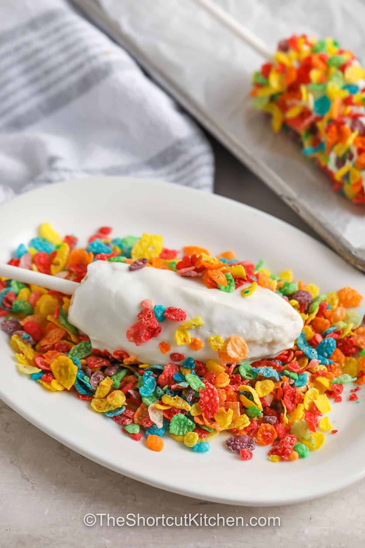 Banana pops make a frozen treat that combines sweet fruit with creamy yogurt and crunchy toppings. This easy recipe is dipped, rolled, and covered with whatever you like, from nuts to chocolate chips. It is a quick, kid-friendly snack that feels like summer in every bite. Keep them in your freezer for a simple way to enjoy a cool snack or dessert whenever you want. Fun to make, even better to eat. #theshortcutkitchen frozen banana dipped in yogurt being rolled in cereal