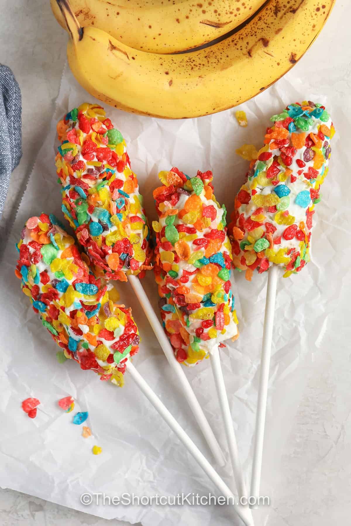 Banana pops are the perfect frozen recipe to enjoy on a warm day. Bananas are dipped in creamy yogurt and covered in crunchy or sweet toppings for a snack that feels like a summer treat. This quick and simple idea is kid-friendly, easy to customize, and delicious to eat. Keep them in your freezer for those times when you want something refreshing, satisfying, and just a little bit sweet. #theshortcutkitchen Four Banana pops coated with fruity cereal