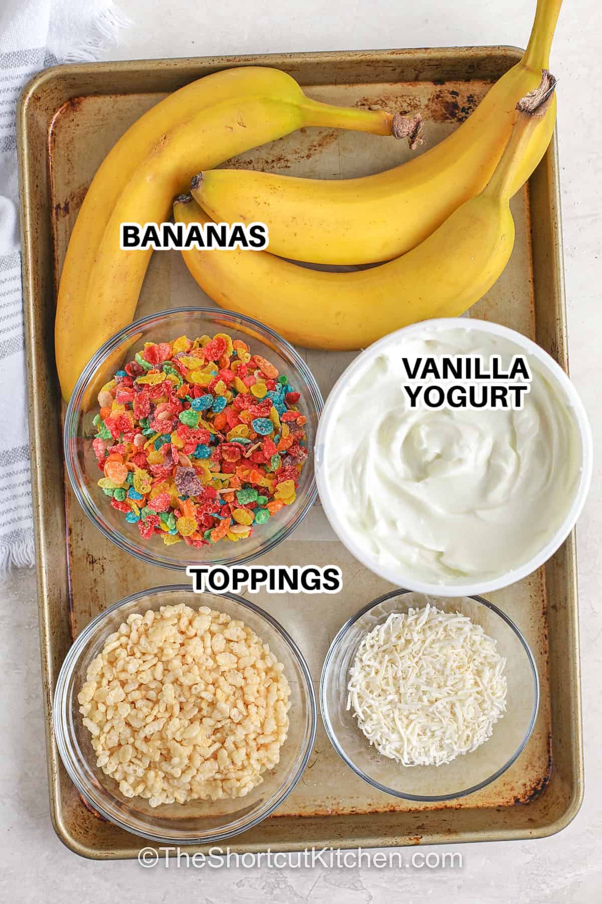 Banana pops are the frozen snack you will love to keep around. This easy recipe has bananas dipped in creamy yogurt and covered with crunchy or sweet toppings for a quick summer treat. They are simple to make, kid-friendly, and fun to enjoy on warm days. You can choose toppings like nuts, coconut, or cereal to make each bite unique. A perfect way to turn fresh bananas into a treat that feels special. #theshortcutkitchen Ingredients to make Banana Pops labeled: bananas, vanilla yogurt, and toppings