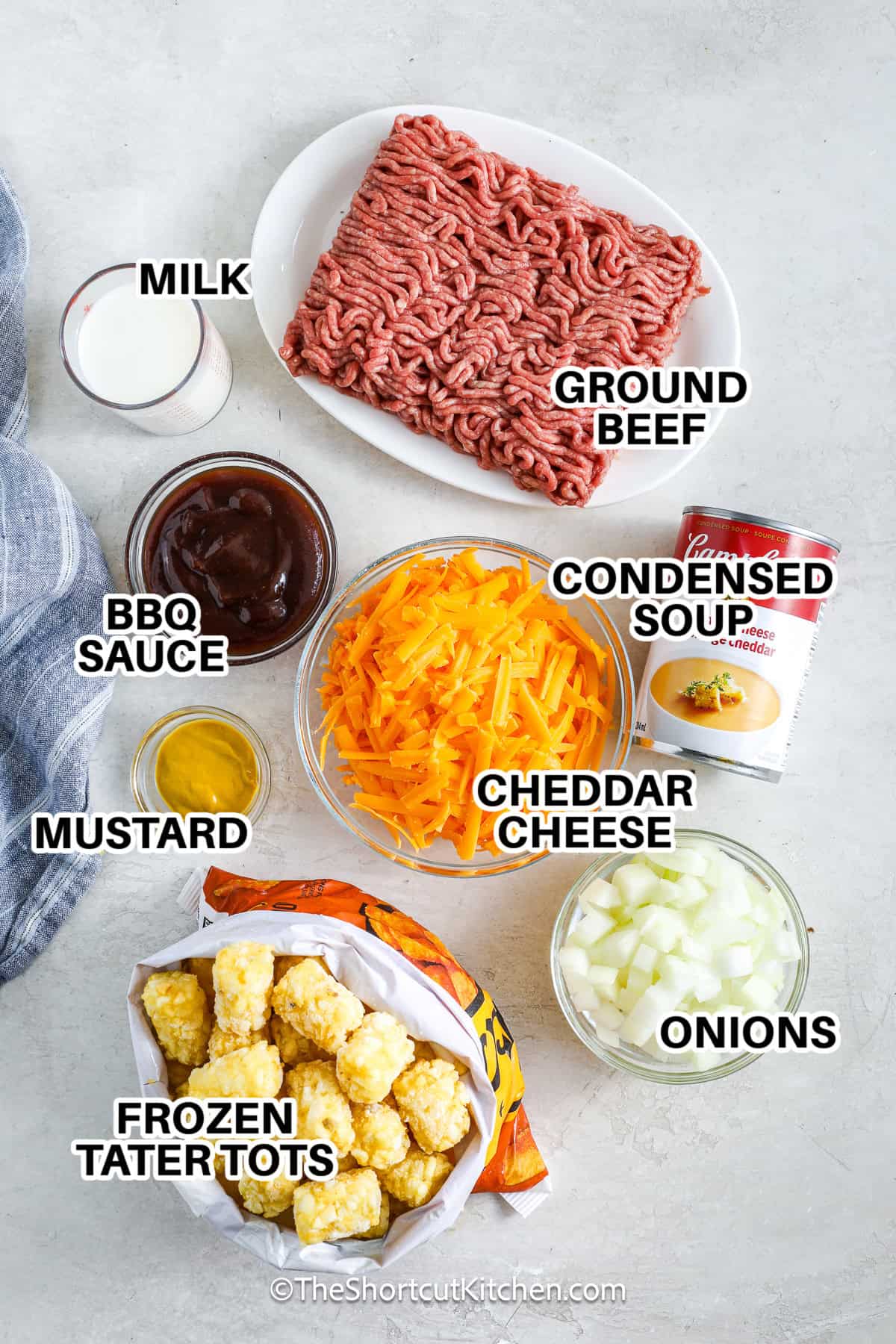 This cheeseburger tater tot casserole is the perfect combination of comfort food and burger-inspired flavor. Savory beef, onions, cheddar soup, mustard, barbecue sauce, and melted cheddar cheese are layered under a crispy tater tot crust and baked until golden and bubbly. Top with chopped pickles or your favorite burger fixings for a fun twist. It’s a hearty, easy, and delicious meal that makes weeknight dinners or family gatherings feel like a special occasion. #theshortcutkitchen Ingredients to make Cheeseburger Tater Tot Casserole labeled: ground beef, milk, bbq sauce, condensed soup, cheddar cheese, onions, mustard, and frozen tater tots