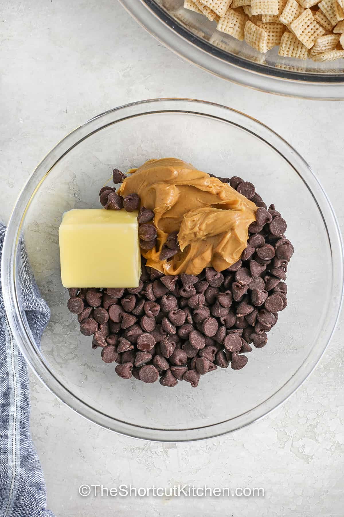 This muddy buddies recipe is a go-to snack for any occasion. This no-bake treat combines Chex cereal with rich chocolate, smooth peanut butter, and a dusting of powdered sugar. Everything comes together in just minutes using the microwave and a large zip-top bag. Perfect for making ahead or as a last-minute dessert when guests are coming. Kids and adults love it. It’s messy, crunchy, and totally irresistible. #theshortcutkitchen A bowl with chocolate chips, butter, and peanut butter to make muddy buddies