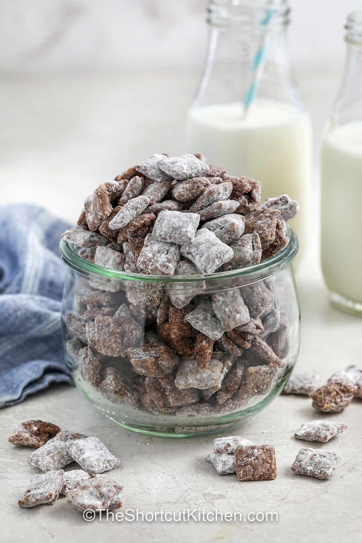 Craving something sweet and crunchy? This muddy buddies recipe hits the spot. Chocolate and peanut butter coat each piece of Chex for a melt-in-your-mouth bite that’s finished with powdered sugar for just the right sweetness. Make it ahead of time and keep it in the freezer for a cold treat on hot days, or enjoy it straight from the bag. This recipe is fast, easy, and perfect for beginners. #theshortcutkitchen a container with prepared muddy buddies