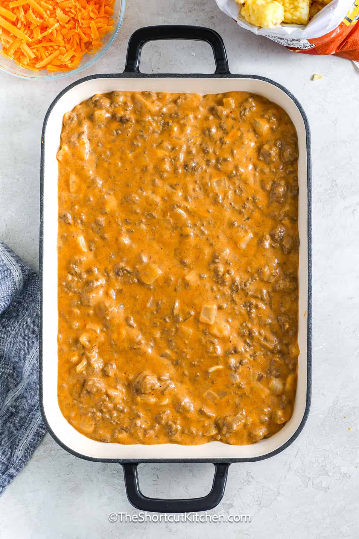 This cheeseburger tater tot casserole is a crowd-pleaser that layers hearty browned beef, creamy cheddar soup mixed with milk and mustard, tangy barbecue sauce, shredded cheddar, and a blanket of tater tots that bake to golden perfection. It’s the ultimate cheesy, savory, and comforting meal that turns simple ingredients into a delicious, indulgent dish everyone will fight over at the table. #theshortcutkitchen cheeseburger tater tot casserole filling in a casserole dish