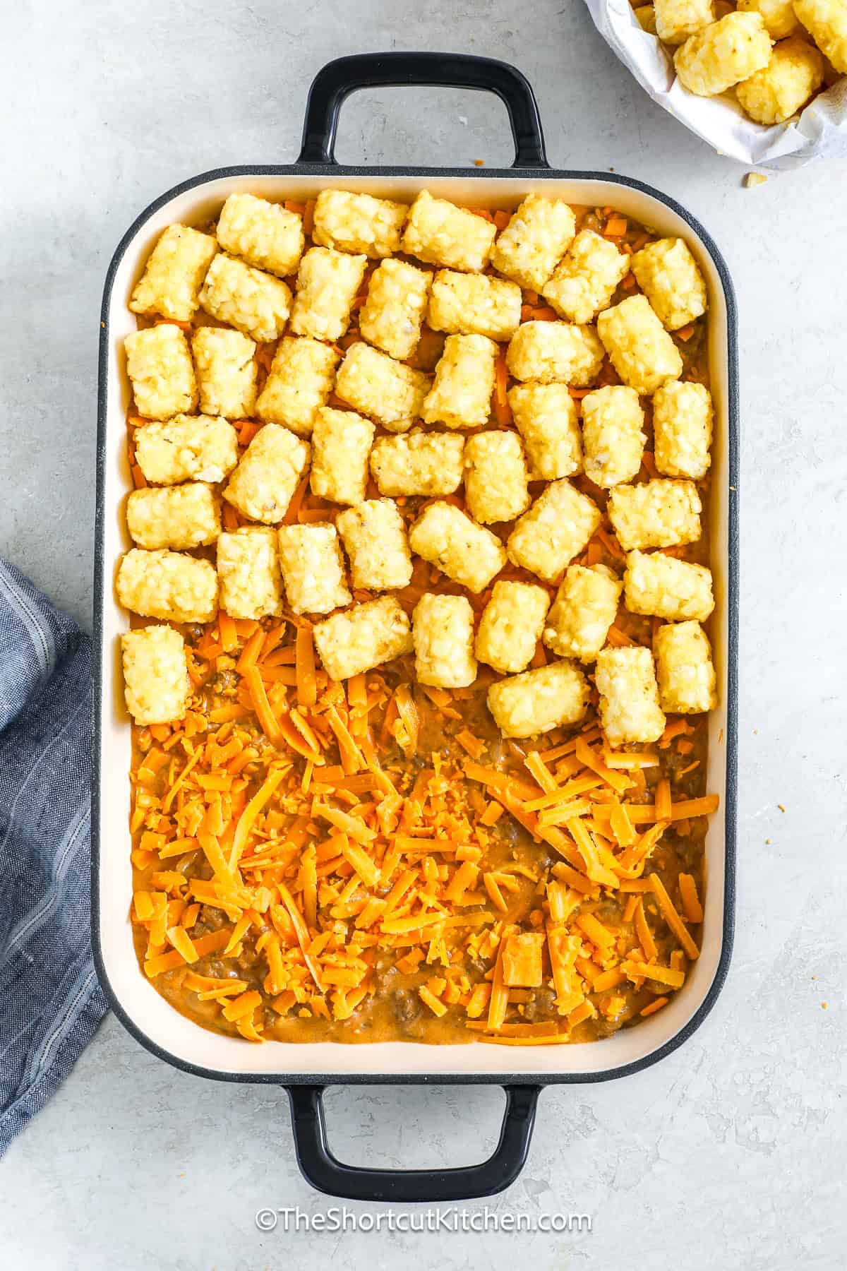 This cheeseburger tater tot casserole is an easy, one-dish dinner that delivers all the flavor of a cheeseburger without flipping patties. Ground beef and onions are simmered with cheddar soup, milk, mustard, and barbecue sauce, then layered with shredded cheddar and topped with tater tots. Baked until bubbly and crisp, it’s perfect for busy weeknights, family dinners, or feeding a hungry crowd with minimal fuss and maximum cheesy satisfaction. #theshortcutkitchen tater tots being added to the cheeseburger tater tot casserole