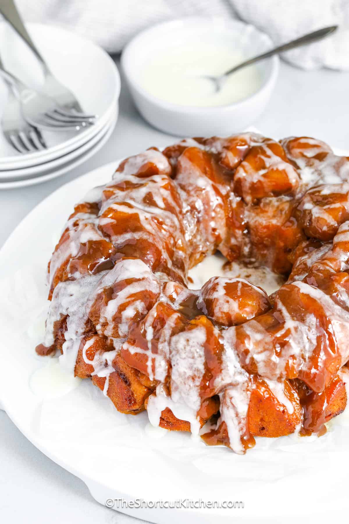 Cinnamon roll monkey bread is a dreamy pull-apart dish that’s easy to make but big on flavor. A rich caramel sauce made from butter, brown sugar, and cream coats every cinnamon roll bite. After you bake it, drizzle with icing for a perfect glazed finish. It’s everything you want in a sweet, gooey, and sticky treat, perfect for holiday brunches, lazy weekend breakfasts, or anytime cravings. This recipe truly is one of the best shortcuts you can use! #theshortcutkitchen Cinnamon Roll Monkey Bread drizzled with icing on a serving plate