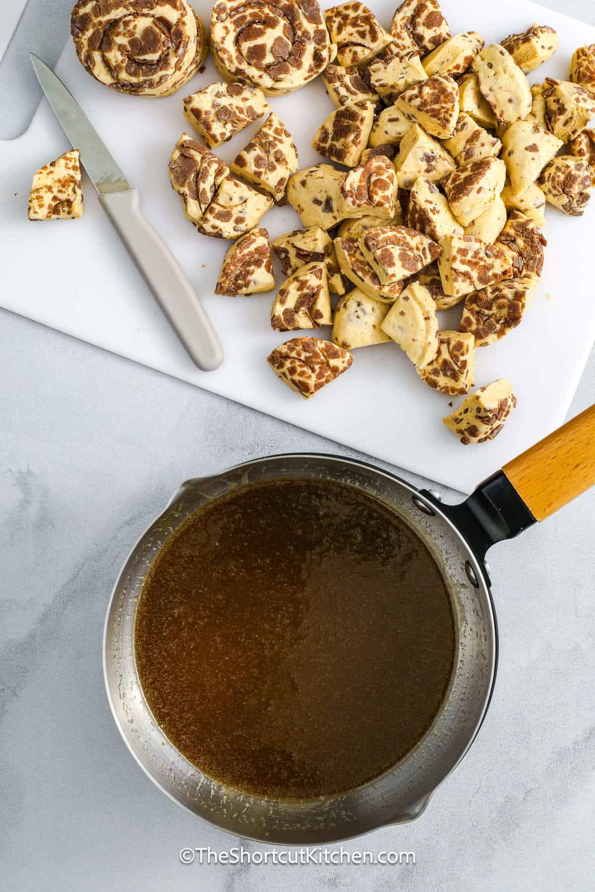 Cinnamon roll monkey bread is a dreamy pull-apart dish that’s easy to make but big on flavor. A rich caramel sauce made from butter, brown sugar, and cream coats every cinnamon roll bite. After you bake it, drizzle with icing for a perfect glazed finish. It’s everything you want in a sweet, gooey, and sticky treat, perfect for holiday brunches, lazy weekend breakfasts, or anytime cravings. This recipe truly is one of the best shortcuts you can use! #theshortcutkitchen monkey bread sauce prepared in a sauce pan and the refrigerated cinnamon rolls chopped on a cutting board