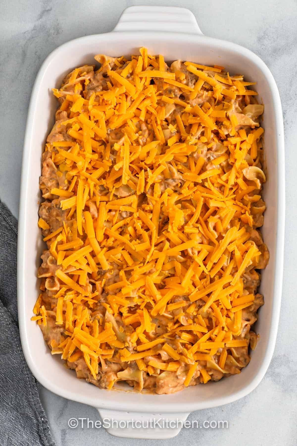 Beef noodle casserole makes dinner simple and satisfying, featuring ground beef, noodles, and a creamy sauce made from mushroom and tomato soups. Sour cream and milk give it a silky texture, while cheddar cheese melts beautifully on top. It’s baked to golden perfection, creating a dish that’s hearty, flavorful, and ready for sharing. Perfect for busy families who love warm, homemade meals. #theshortcutkitchen adding cheese on top of Beef Noodle Casserole before cooking