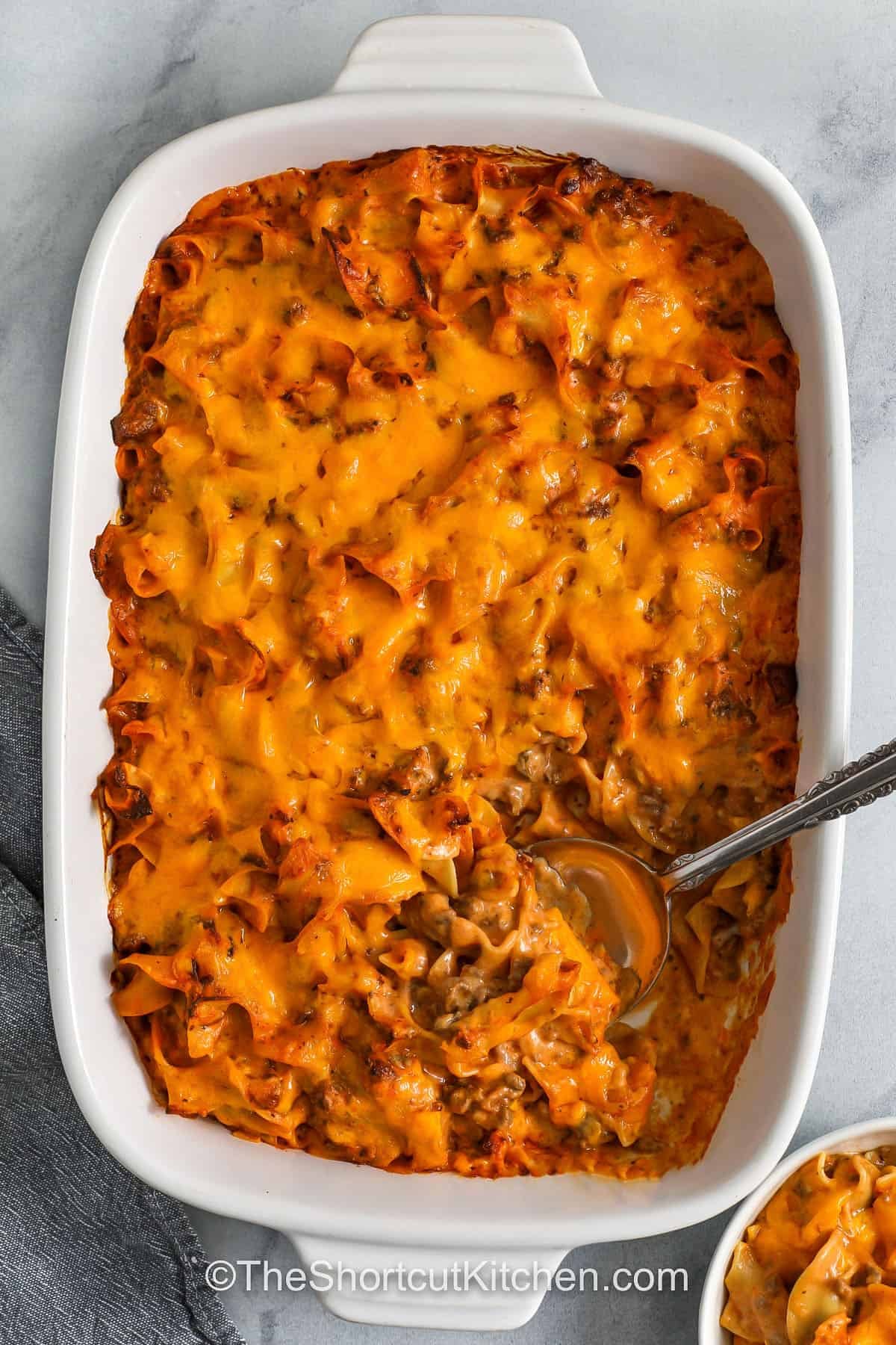 Beef noodle casserole delivers everything you love about classic comfort food. It has a creamy texture, rich flavors, and gooey cheese. With tender noodles, savory beef, and a blend of mushroom and tomato soups, it’s both simple and delicious. Baked to perfection with melted cheddar, this hearty meal is ideal for busy nights or feeding a hungry family. It’s cozy, filling, and oh-so-satisfying. #theshortcutkitchen Beef Noodle Casserole in a casserole dish with a part taken out