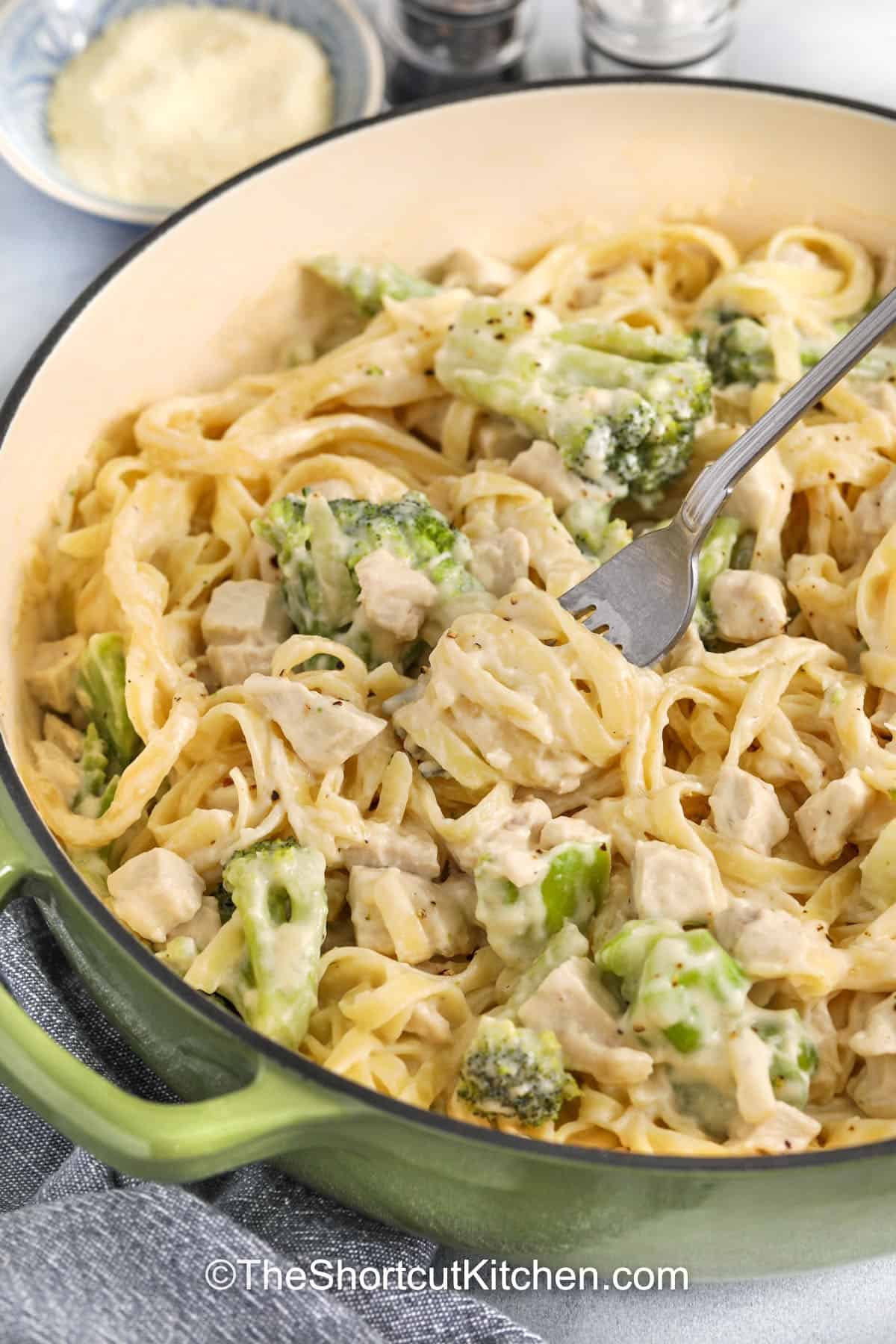 Chicken Alfredo with broccoli is the ultimate comfort dinner, combining tender fettuccine pasta, juicy chicken, and crisp broccoli florets in a rich, creamy, cheesy sauce. This is ready in just minutes. Perfect for busy weeknights, it’s family-friendly, indulgent, and satisfying without the stress of complicated cooking. Everyone at the table will love it. #theshortcutkitchen pot of Chicken Alfredo with Broccoli with a fork