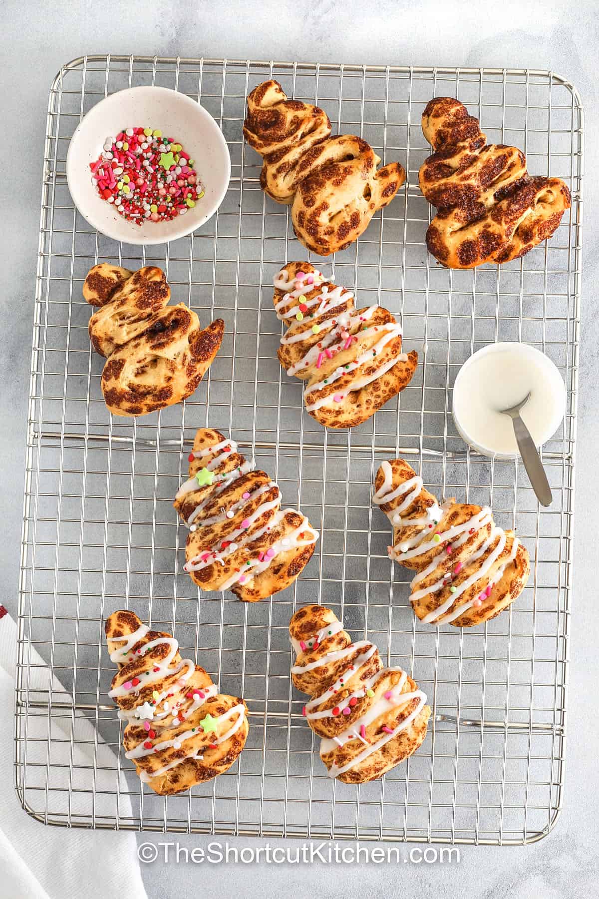 Cinnamon roll Christmas tree treats turn ordinary cinnamon rolls into a festive holiday masterpiece. Easy to assemble and bake, they are soft, fluffy, and packed with sweet, warm cinnamon flavor. Decorated with icing and colorful sprinkles, these adorable pastries are a fun and easy way to delight kids and adults alike during the holiday season. #theshortcutkitchen adding icing to Cinnamon Roll Christmas Tree