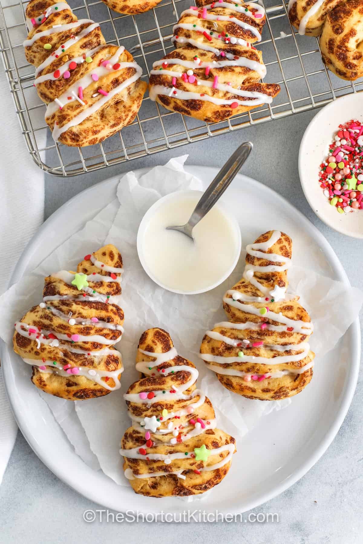 Cinnamon roll Christmas tree creations are a delightful way to celebrate the season with family. Each tree-shaped roll is tender, buttery, and filled with warm cinnamon-sugar goodness, then topped with icing and festive sprinkles. Quick to bake and fun to decorate, they make a cheerful centerpiece for your breakfast or brunch table while keeping the holiday spirit sweet and simple. #theshortcutkitchen plated Cinnamon Roll Christmas Tree with icing