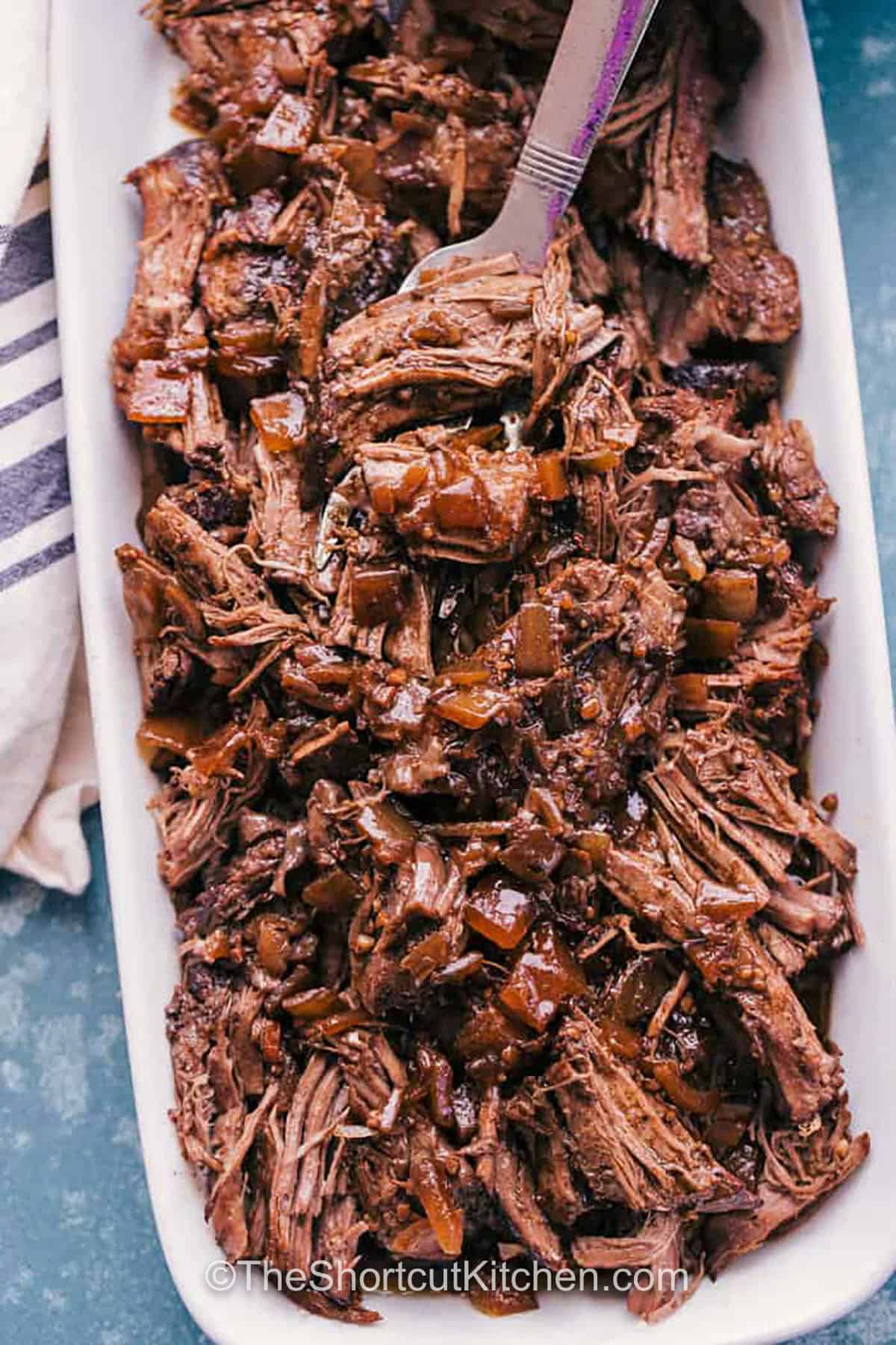 This Crockpot roast beef recipe uses a mix of broth, balsamic glaze, honey, garlic, and onions to turn a simple chuck or round roast into chunks of tender beef filled with flavor for any family dinner or holiday table. When the roast is shredded and topped with warm juices, the flavor becomes rich and balanced, making it an easy favorite. A spoonful of creamy mashed potatoes completes the meal in the best way. #theshortcutkitchen juicy Crockpot Roast Beef Recipe on a plate
