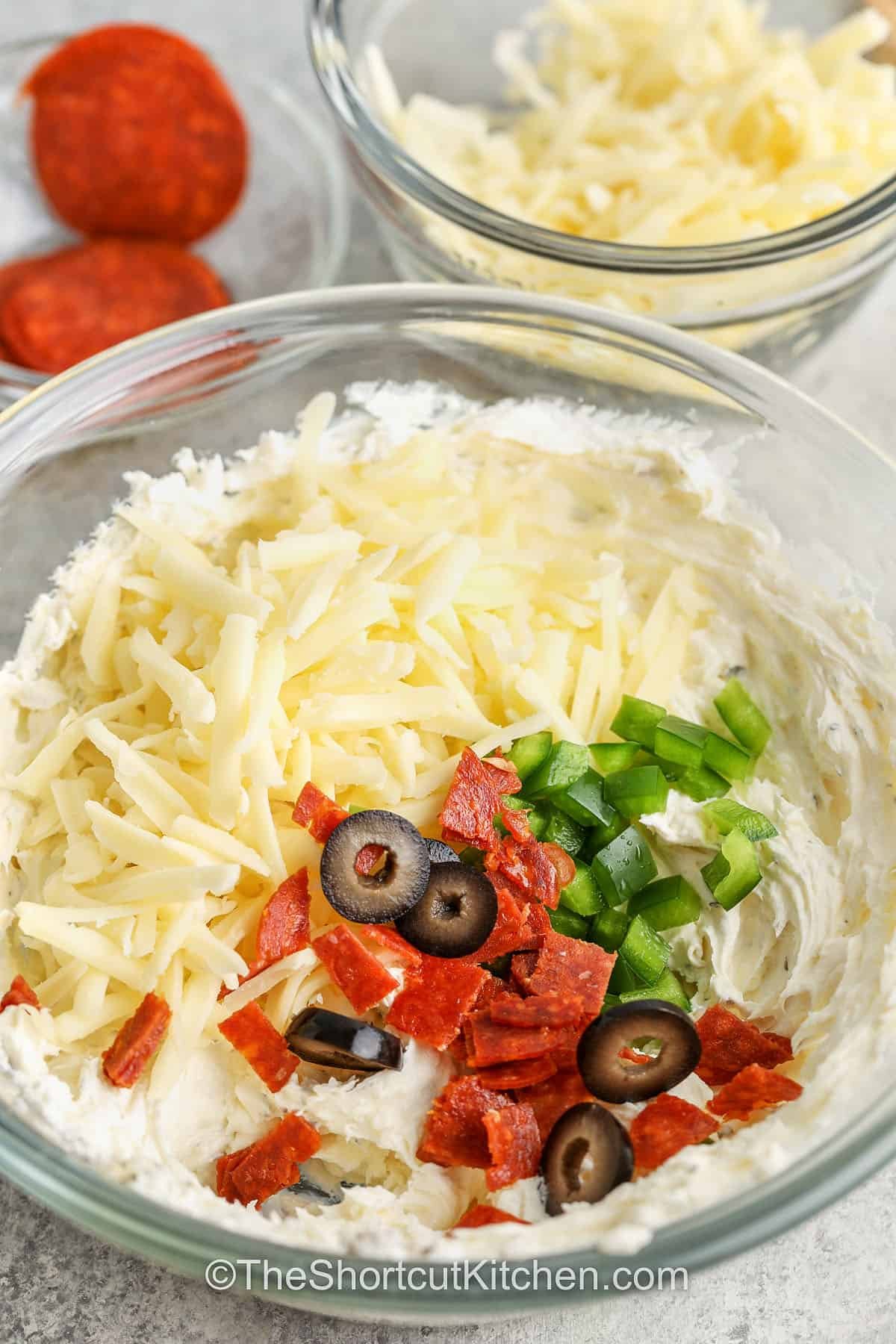 This pizza dip recipe is a creamy, cheesy way to make any night special. With gooey mozzarella, garlic, sauce, and pepperoni, it’s everything you love about pizza in dip form. The hot, melty layers make it a crowd favorite every time. It’s so easy to make, and the best part is watching everyone scoop it up fast. Serve with chips or bread for the ultimate party snack. #theshortcutkitchen adding cheese and toppings to cream cheese mixture to make Pizza Dip Recipe