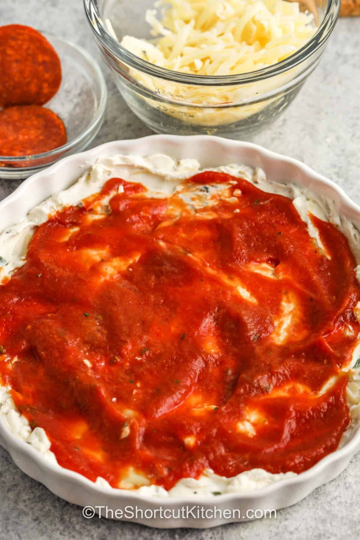 This pizza dip recipe makes it easy to bring the best flavors of pizza to your snack table. Creamy cheese, tangy sauce, and garlic blend for a rich, hot dip that’s full of pepperoni goodness. It’s the kind of easy appetizer everyone asks for again and again. Make it for movie night, parties, or a quick weekend treat that’s simple and satisfying to the last bite. #theshortcutkitchen adding pizza sauce on top of cream cheese mixture to make Pizza Dip Recipe