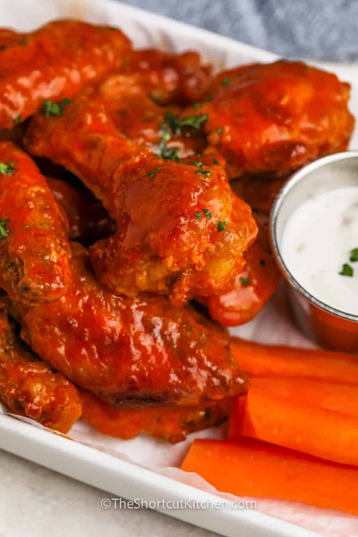 Love wings but hate the deep frying? Super crispy baked buffalo wings are the answer. Shake wings with flour, baking powder, garlic, and cayenne, chill to dry, then bake until crispy and cooked through. Finish with buttery Frank’s hot sauce for that bold buffalo flavor. These crispy baked buffalo wings are perfect for appetizers, snack boards, or a casual meal with fries and dip. #theshortcutkitchen baked buffalo wings on a platter with dip and carrot sticks