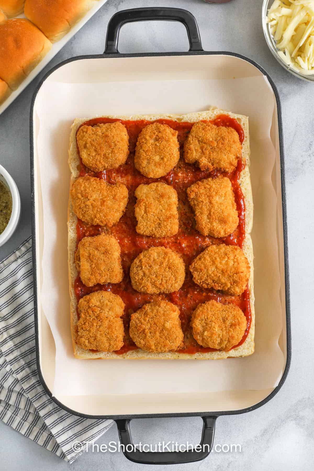 Chicken parm sliders made the shortcut way. Use frozen chicken tenders (cut to fit) or nuggets, add marinara, mozzarella, and parmesan, then bake on Hawaiian rolls with a garlic butter topping. These chicken parmesan sliders come out melty and golden, perfect for feeding a group. Serve with extra marinara for dipping and a simple salad on the side. #theshortcutkitchen chicken parm sliders with chicken nuggets in a pan