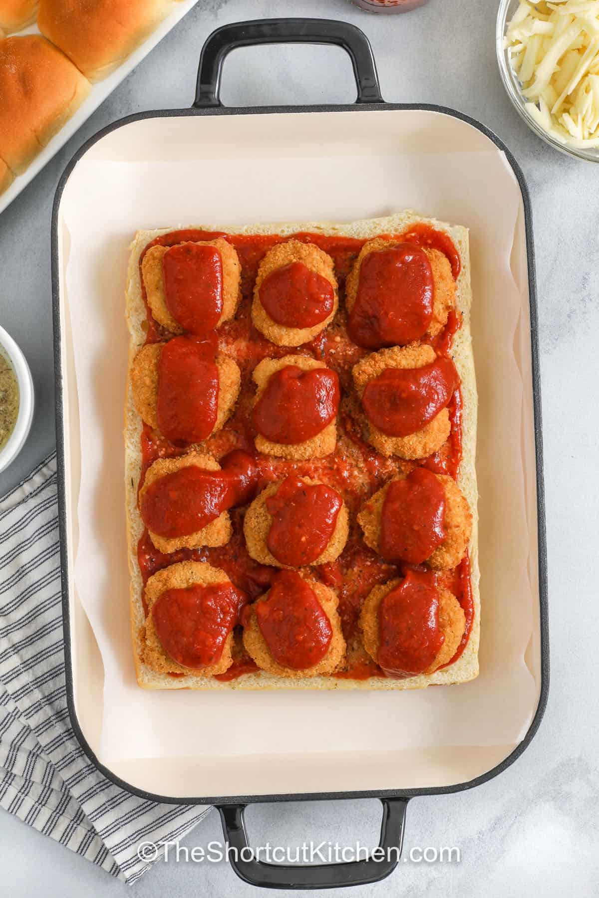 Turn freezer staples into chicken parm sliders everyone loves. Bake frozen nuggets, layer with marinara sauce and mozzarella on Hawaiian rolls, sprinkle parmesan, and brush with garlic butter before baking. These baked chicken parmesan sliders are cheesy and dip-worthy, with a sweet-savory roll base and a crisp, buttery top. Great for quick dinners and game day snacks. #theshortcutkitchen chicken parm sliders with marinara in a pan