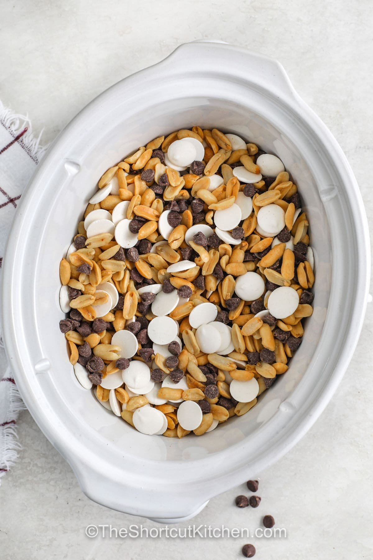 Make Christmas Crockpot peanut clusters with just three simple ingredients. Chocolate chips and almond bark melt in the slow cooker before coating roasted peanuts for the easiest holiday candy. This recipe is great for gifting, parties, or storing for last-minute Christmas treats. #theshortcutkitchen adding ingredients to crockpot to make Crockpot Peanut Clusters