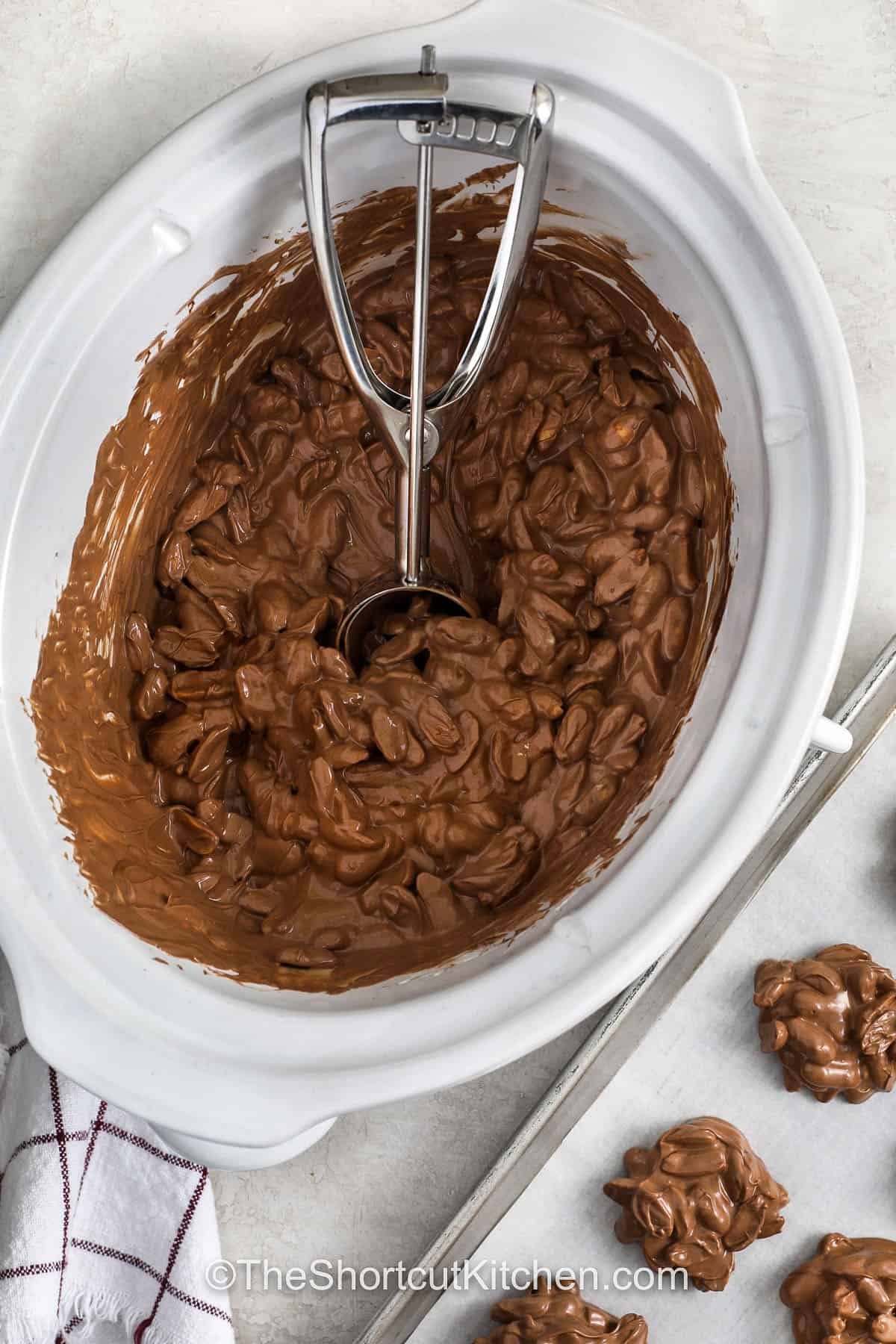 Easy Crockpot peanut clusters are perfect for Christmas candy trays. The slow cooker melts semisweet chocolate and almond bark into a creamy mixture that coats roasted peanuts with sweet-salty flavor. These clusters require almost no effort, make a big batch, and freeze well for holiday prep. #theshortcutkitchen scooping Crockpot Peanut Clusters from the pot to a sheet pan