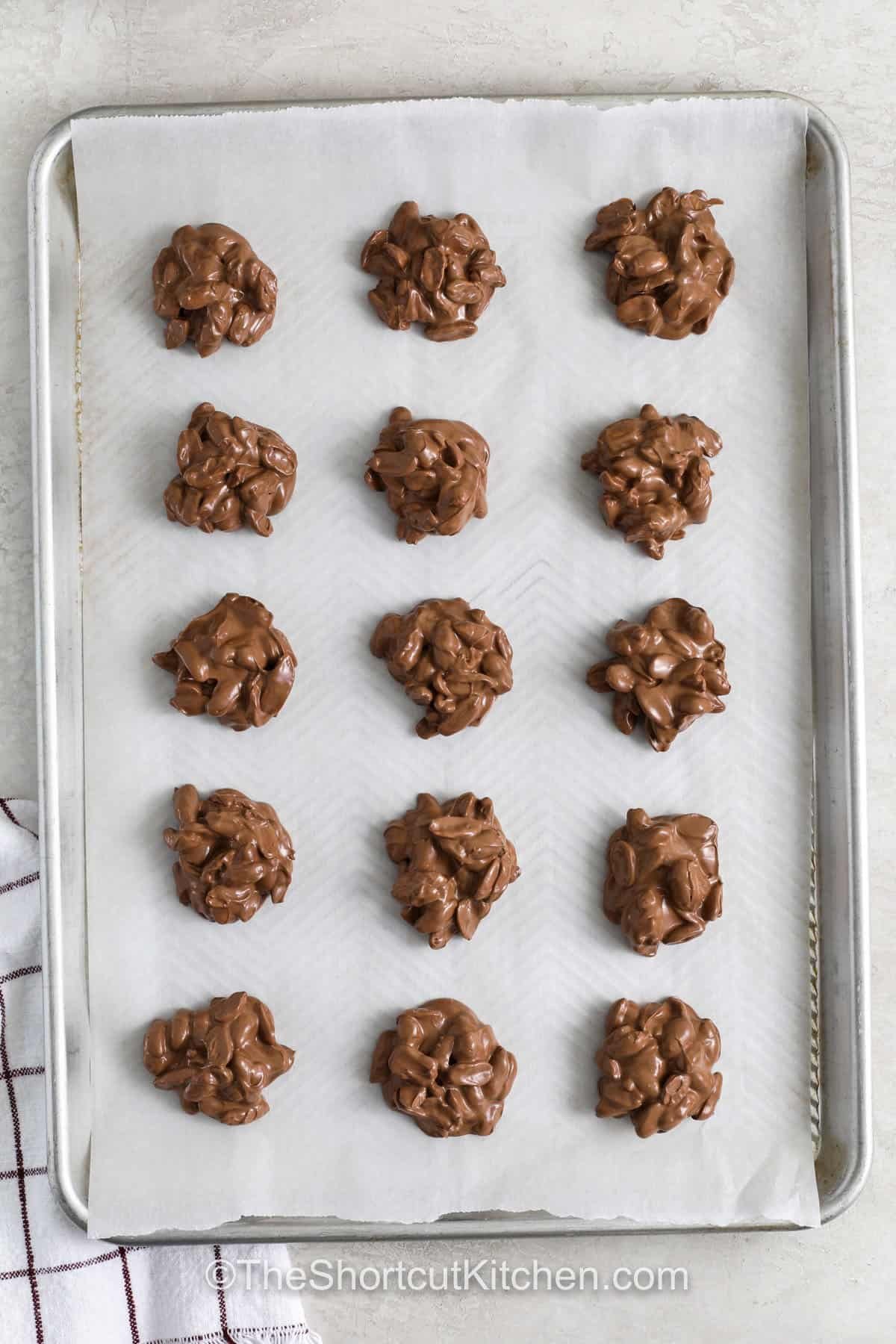 Crockpot peanut clusters make a cozy Christmas candy that’s sweet, crunchy, and easy to prepare. Almond bark and chocolate chips melt together into a smooth coating for roasted peanuts. Drop spoonfuls on parchment and let cool for a festive treat that works for parties, gifting, or stocking holiday dessert boards. #theshortcutkitchen sheet pan of Crockpot Peanut Clusters