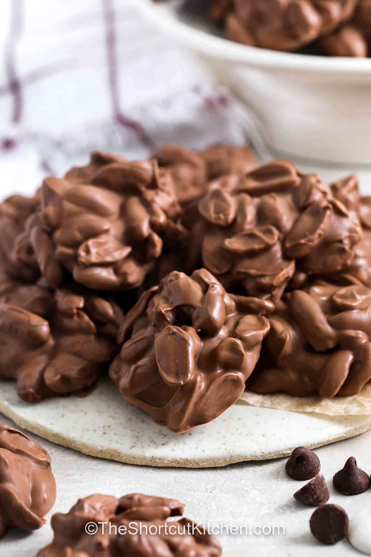 This Crockpot peanut clusters recipe creates delicious Christmas candy with minimal work. The slow cooker does the melting, and slightly salted roasted peanuts add the perfect crunch. Scoop and cool for sweet-salty treats that store well and make great gifts. With big-batch yield and simple ingredients, this recipe is perfect for any holiday celebration. #theshortcutkitchen plate of Crockpot Peanut Clusters