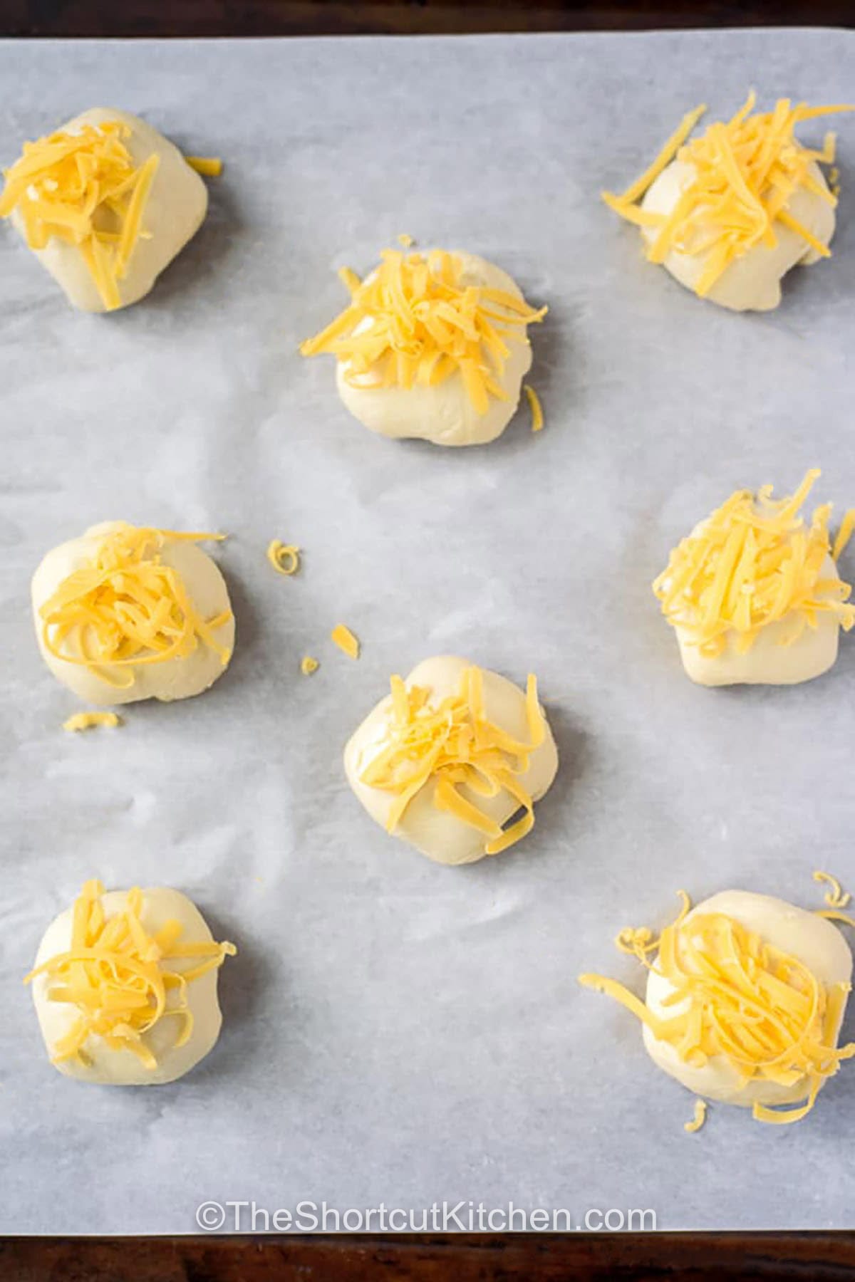 Bacon cream cheese biscuits are cheesy bacon biscuit bombs made with shortcuts. Stuff biscuit dough with a creamy blend of bacon, cream cheese, parmesan, green onion, and garlic powder, then bake until puffed and golden with cheddar on top. Serve warm with soup, chili, or as a party bite. Avoid overfilling so the seams stay sealed and tidy. #theshortkitchen adding cheese to Bacon Cream Cheese Biscuits before baking