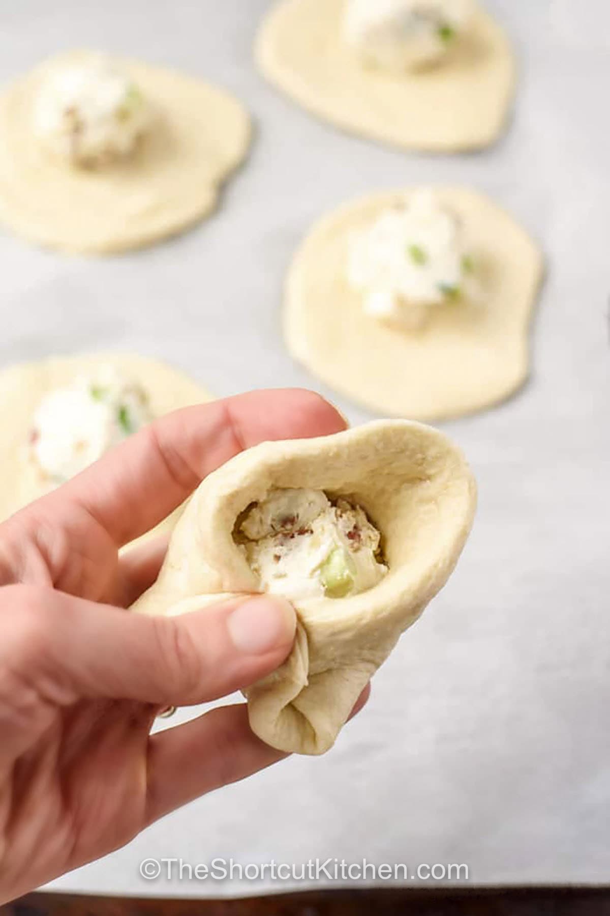 Need a fast appetizer that feels special. Bacon cream cheese biscuits deliver. Flaky refrigerated biscuits are filled with a creamy bacon-cheese mixture, pinched closed, and baked with cheddar on top. Think biscuit bombs with a warm, melty center. Perfect for game day spreads, brunch, or an easy make-ahead snack. Freeze baked biscuits and reheat for busy mornings. #theshortkitchen folding biscuit dough over filling to make Bacon Cream Cheese Biscuits