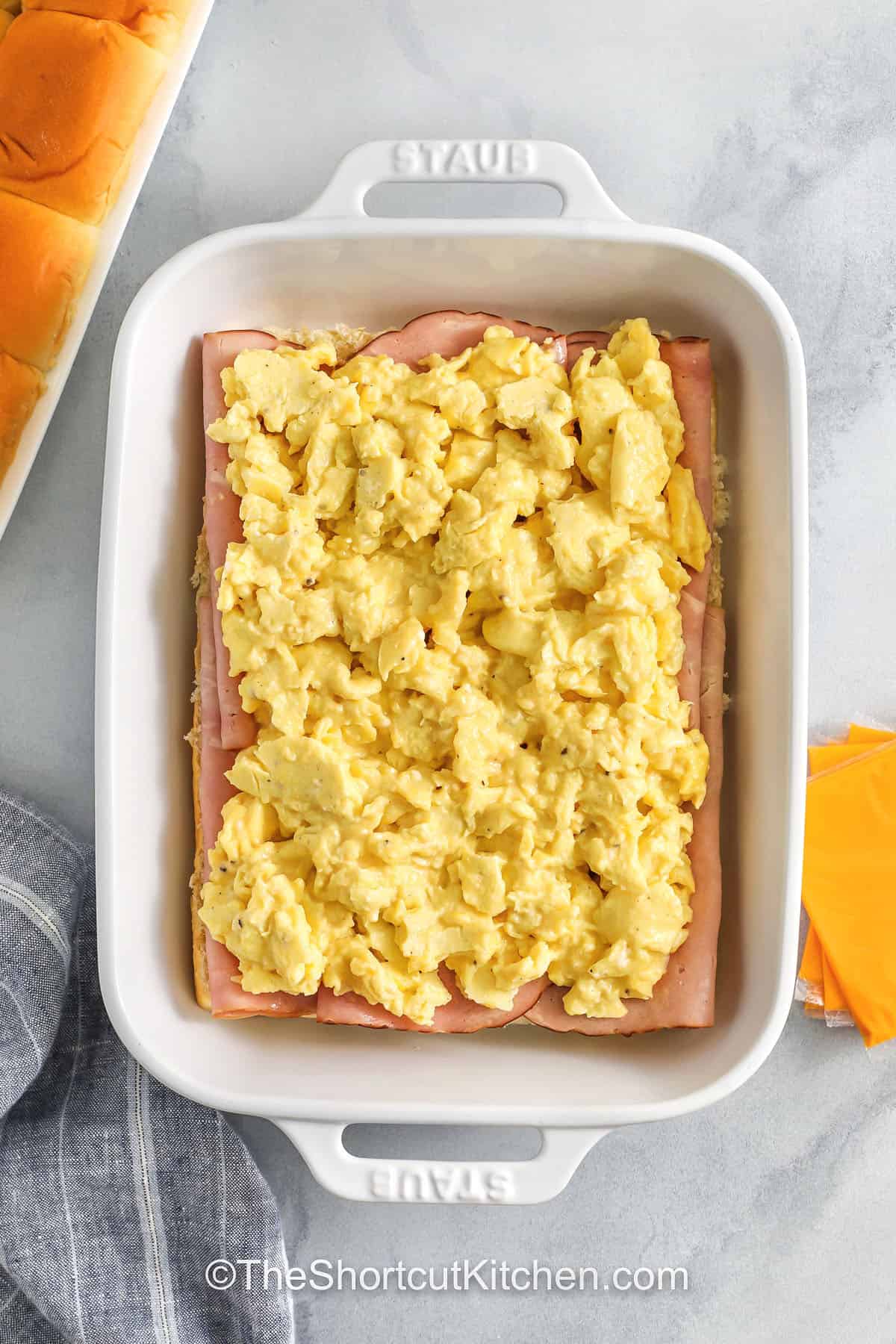 One pan breakfast sliders with Hawaiian rolls are perfect for meal prep. Assemble with scrambled eggs, ham, and melty Swiss or American cheese, then bake until warmed through. Finish with a buttery everything bagel seasoning glaze for flavor-packed tops. Slice into 12 sliders for grab-and-go breakfasts that reheat beautifully in the oven or air fryer. #theshortcutkitchen adding eggs to dish to make Breakfast Sliders