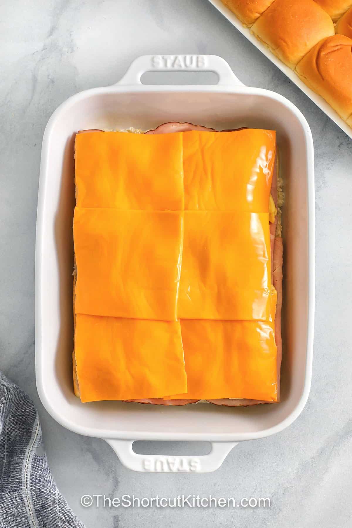 Ham egg and cheese breakfast sliders are a cozy, satisfying breakfast that slices perfectly. Keep rolls attached, soft-scramble the eggs, and layer everything in a pan. The buttery everything bagel seasoning topping with a touch of honey makes the tops golden and irresistible. Great for make ahead breakfasts, holiday mornings, and feeding hungry teens. #theshortcutkitchen adding cheese slices to dish to make Breakfast Sliders