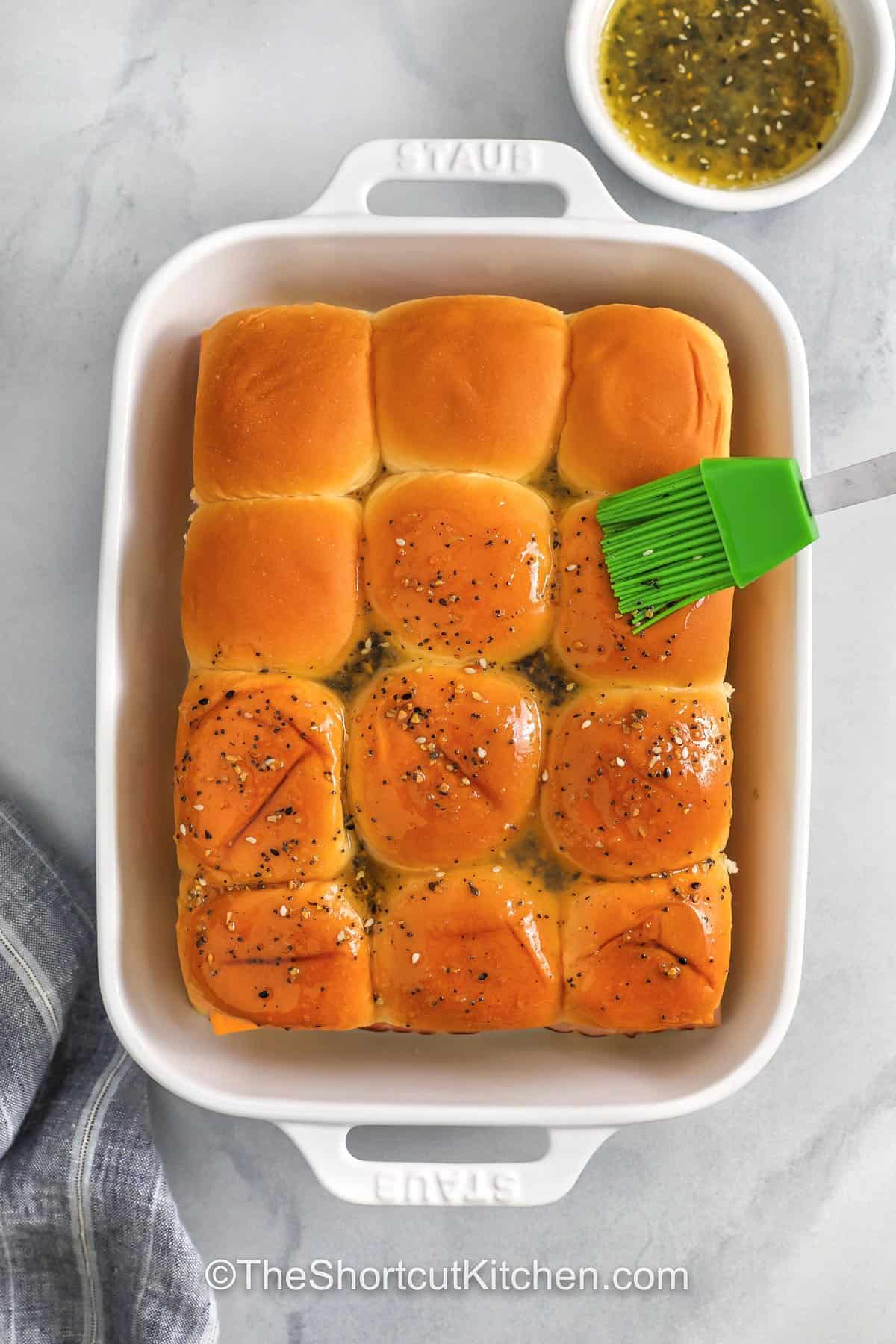 Breakfast sliders make brunch effortless. Use Hawaiian rolls, Egg Beaters or eggs, deli ham, and cheddar for a quick assembly, then bake covered and uncover to lightly brown. The everything bagel seasoning butter topping adds crunch and flavor without extra work. Serve with fruit or home fries, and store leftovers for easy reheating. #theshortcutkitchen spreading butter over Breakfast Sliders in dish