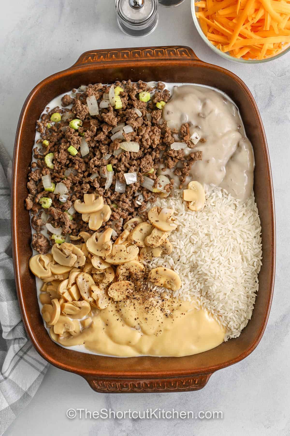 This cheesy hamburger casserole is a total dinner win. Hamburger rice casserole combines browned ground beef with onion and celery, then mixes with uncooked rice, condensed cream soups, and milk for a creamy, cozy bake. Sprinkle shredded cheddar cheese on top to finish. It’s an easy weeknight casserole that’s budget-friendly, family-size, and simple to customize with veggies. #theshortcutkitchen ingredients in casserole dish to make Hamburger Rice Casserole