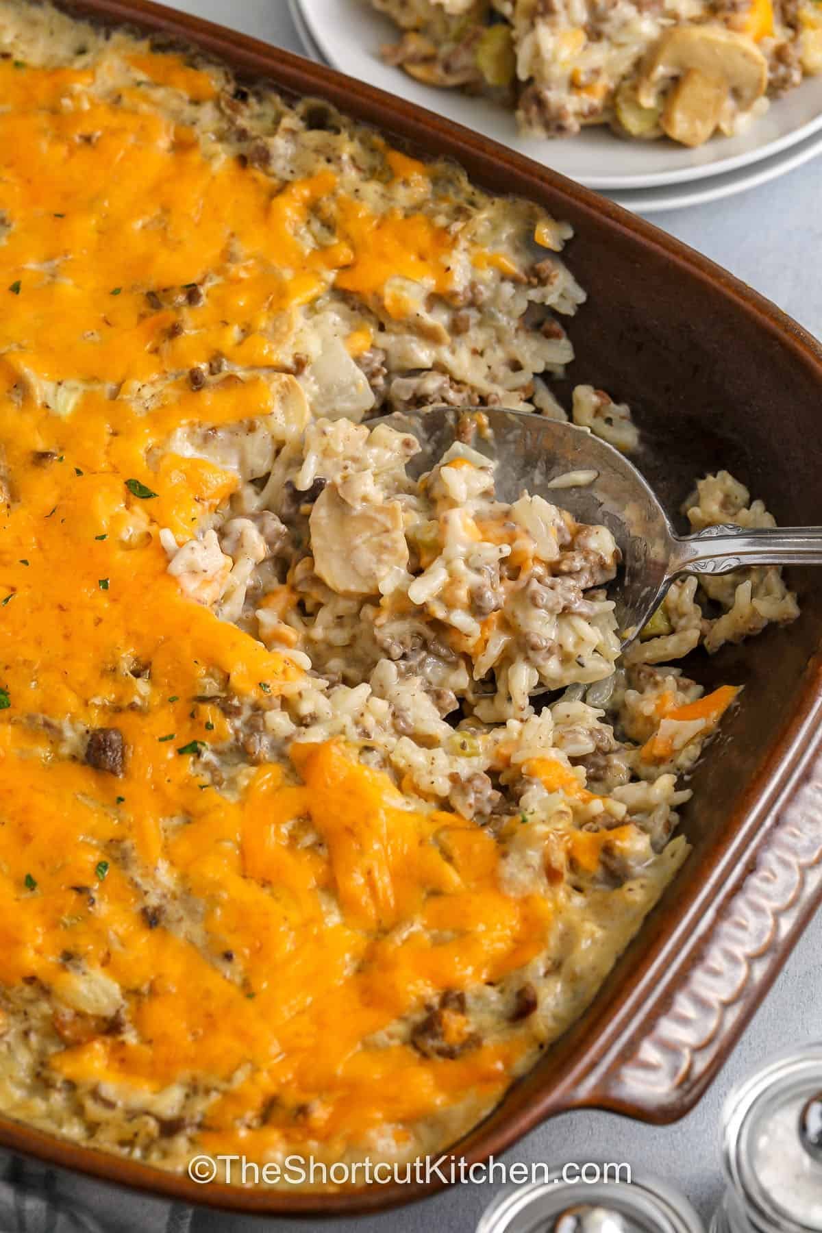 Need an easy weeknight casserole that feeds a crowd. Hamburger rice casserole is a creamy ground beef and rice casserole made with pantry condensed soups, milk, and uncooked rice that bakes up tender and comforting. Top with cheddar cheese for a bubbly, cheesy hamburger casserole everyone loves. Add mushrooms if you like, or swap cream of celery for a milder flavor. #theshortcutkitchen taking a spoonfull of Hamburger Rice Casserole out the casserole dish