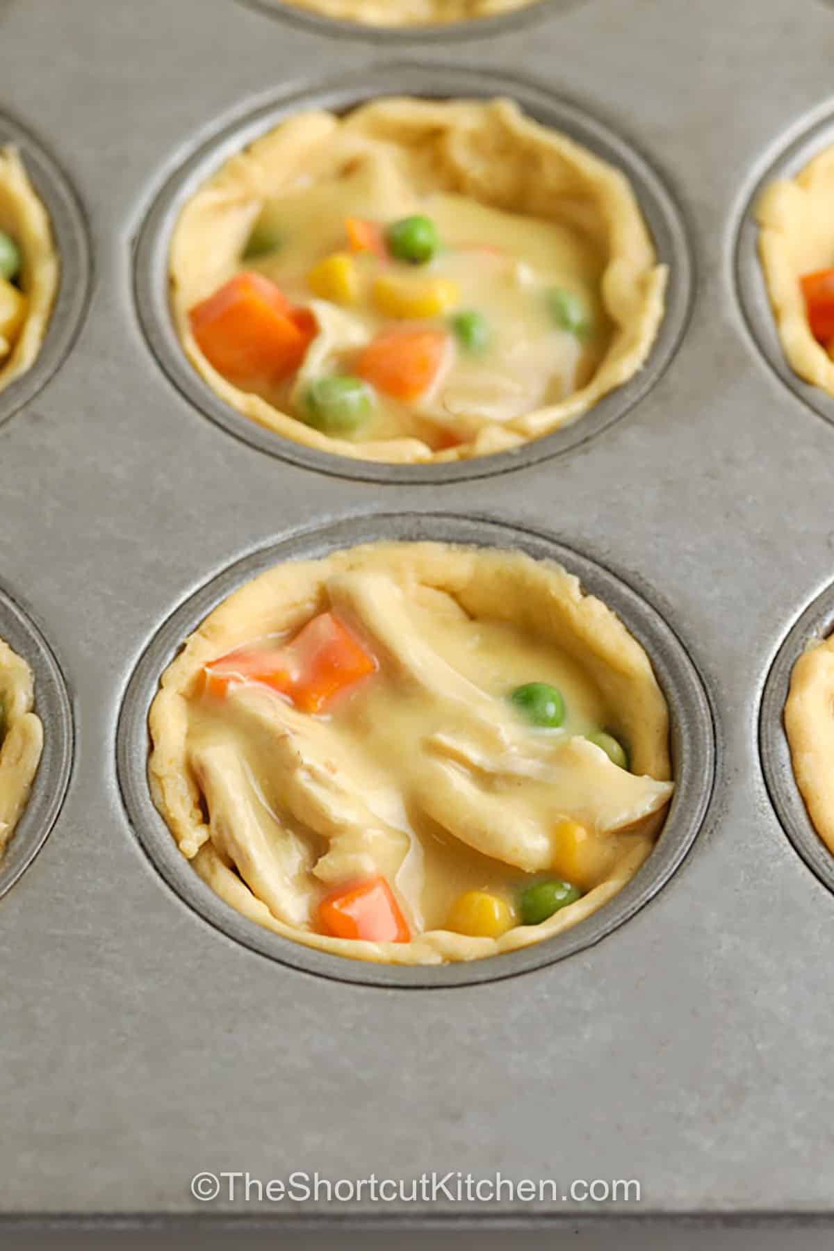 Crescent roll mini chicken pot pies are the ultimate shortcut recipe. A creamy sauce made from condensed cream of chicken soup coats chopped cooked chicken and mixed veggies, then everything bakes in flaky crescent cups. These muffin tin chicken pot pies are great for quick dinners, potlucks, and game day. Make ahead by prepping the filling early and baking right before serving. #theshortcutkitchen Mini Chicken Pot Pies in muffin tin before cooking