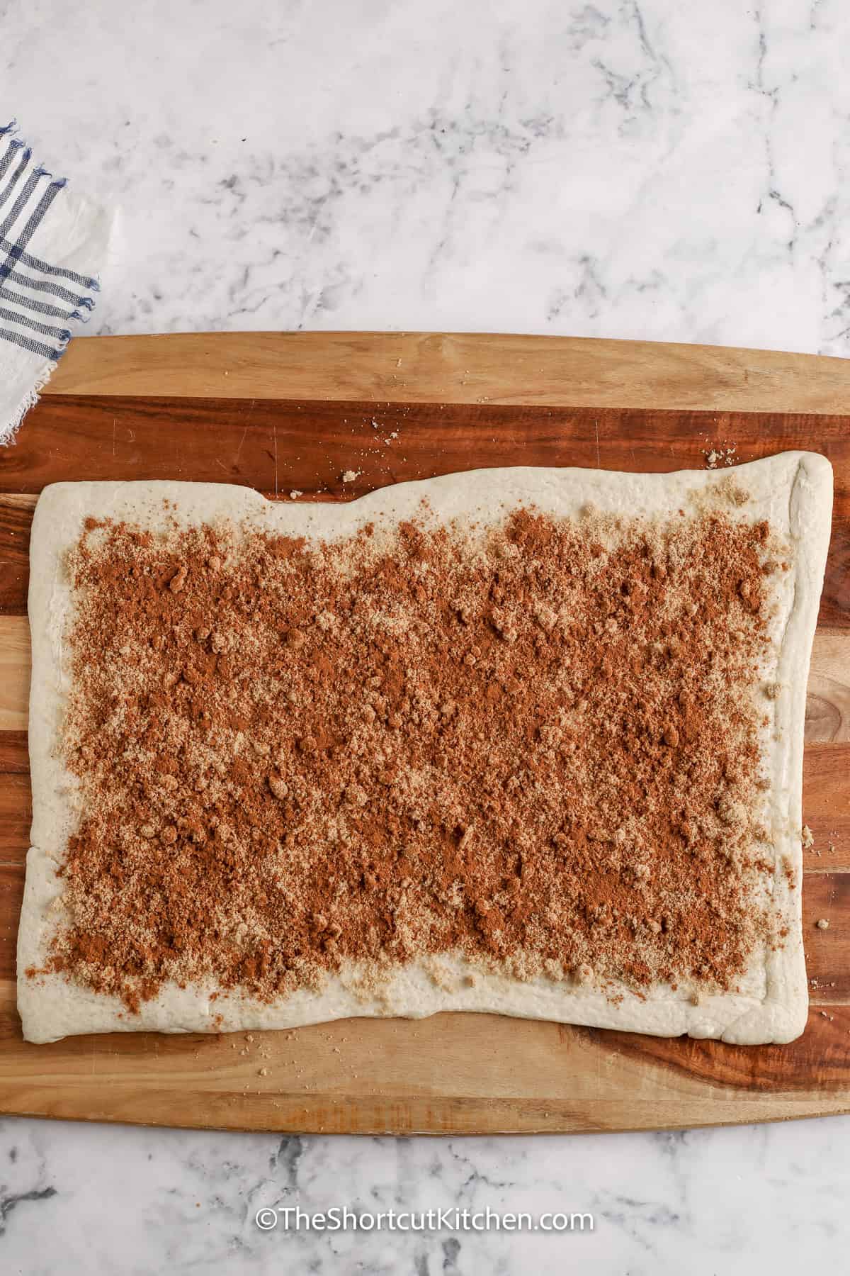 These easy cinnamon rolls are made with pizza dough, and they taste like a coz,y homemade weekend treat. Roll refrigerated dough into a rectangle, add softened butter, cinnamon, and brown sugar, then just roll, slice, and bake. Drizzle a quick cinnamon roll glaze while they are warm. Try a crescent roll version for a flaky cinnamon roll that’s easy and quick to make. #theshortcutkitchen adding brown sugar and cinnamon to pizza dough to make Pizza Dough Cinnamon Rolls